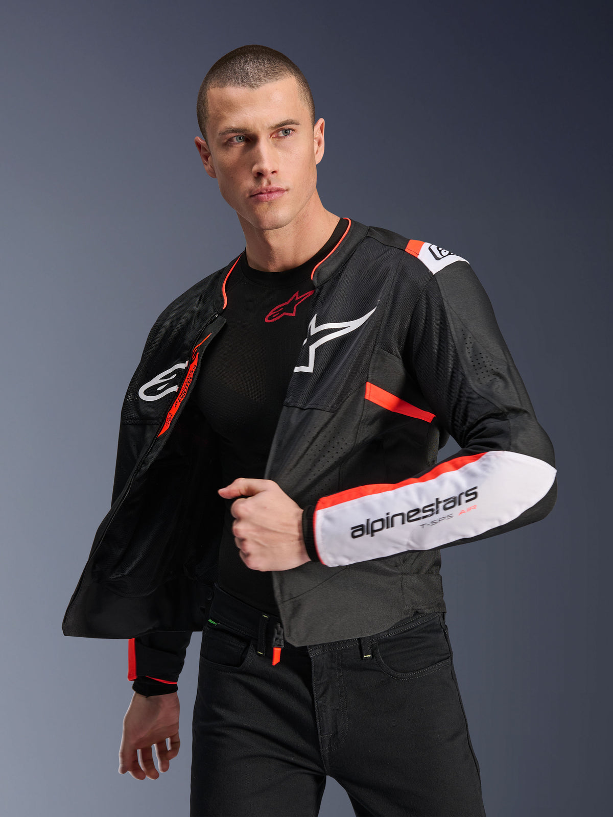 Alpinestars T-SPS Air V2 Jacke, schwarze Textil-Motorradjacke mit weißen und fluoreszierend roten Akzenten, Mesh-Einsätze für Luftzirkulation, weißes Alpinestars-Logo auf der Brust und Branding am Ärmel, männliches Modell trägt die Jacke offen über einem schwarzen Baselayer
