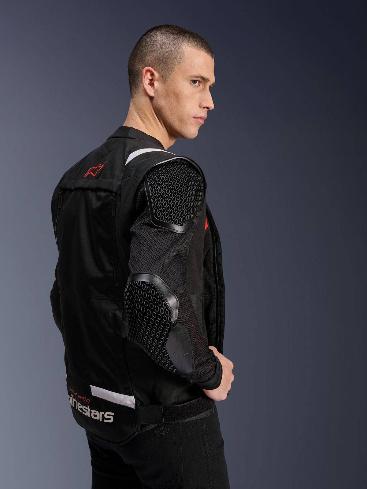 Alpinestars T-Stunt Air Jacke, Motorradjacke, Schwarz, Seitenprofil eines Models, das die sportlich geschnittene Jacke mit Mesh-Ärmeln und sichtbaren Protektoren mit Wabenmuster an Schulter und Ellbogen trägt, rotes Logo auf der Rückseite, weißes Branding an der Taille.