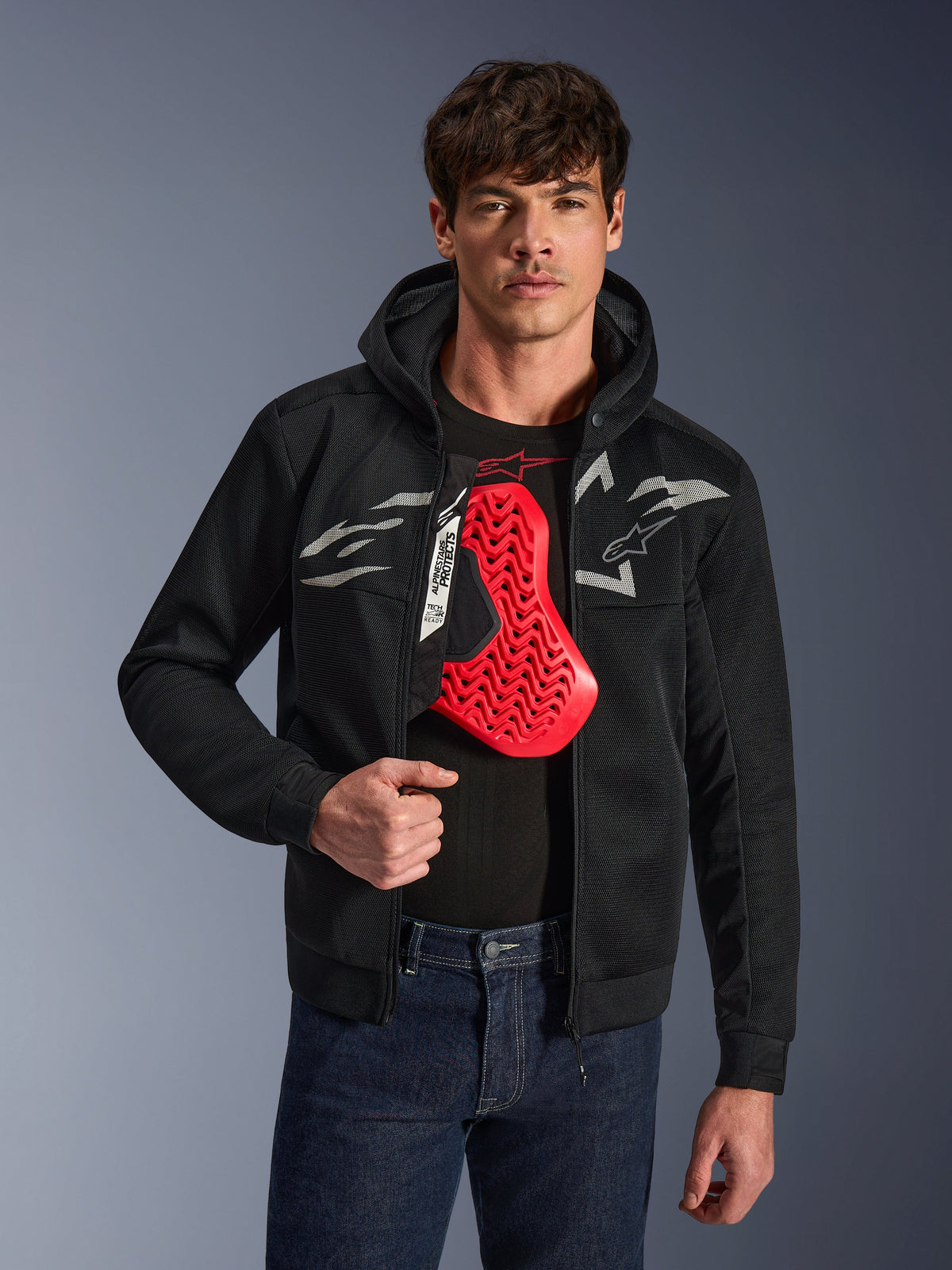 Alpinestars Chrome Superairflow Hoodie in Black, featuring white and gray Astars logos, worn by a male model with a red chest protector visible underneath, 3D mesh textile for ventilation, sporty urban motorcycle apparel.