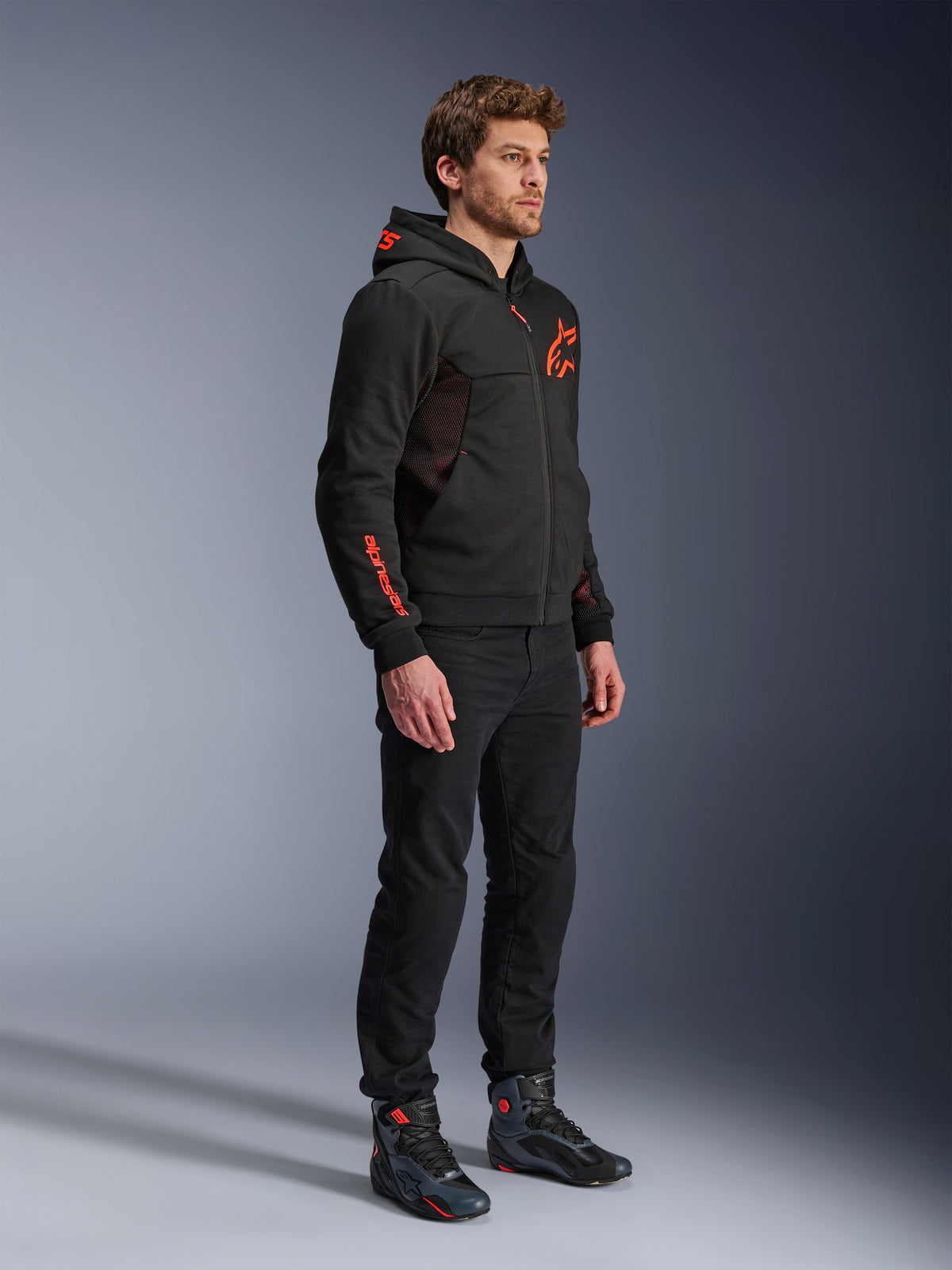 Alpinestars Chrome Air Sport Hoodie, Schwarz Rot Fluo, urbaner Motorrad-Hoodie aus Tech-Fleece, kombiniert mit schwarzer Hose und Motorradstiefeln, mit Mesh-Belüftungseinsätzen und fluoreszierend roten Logos auf Brust und Ärmel.