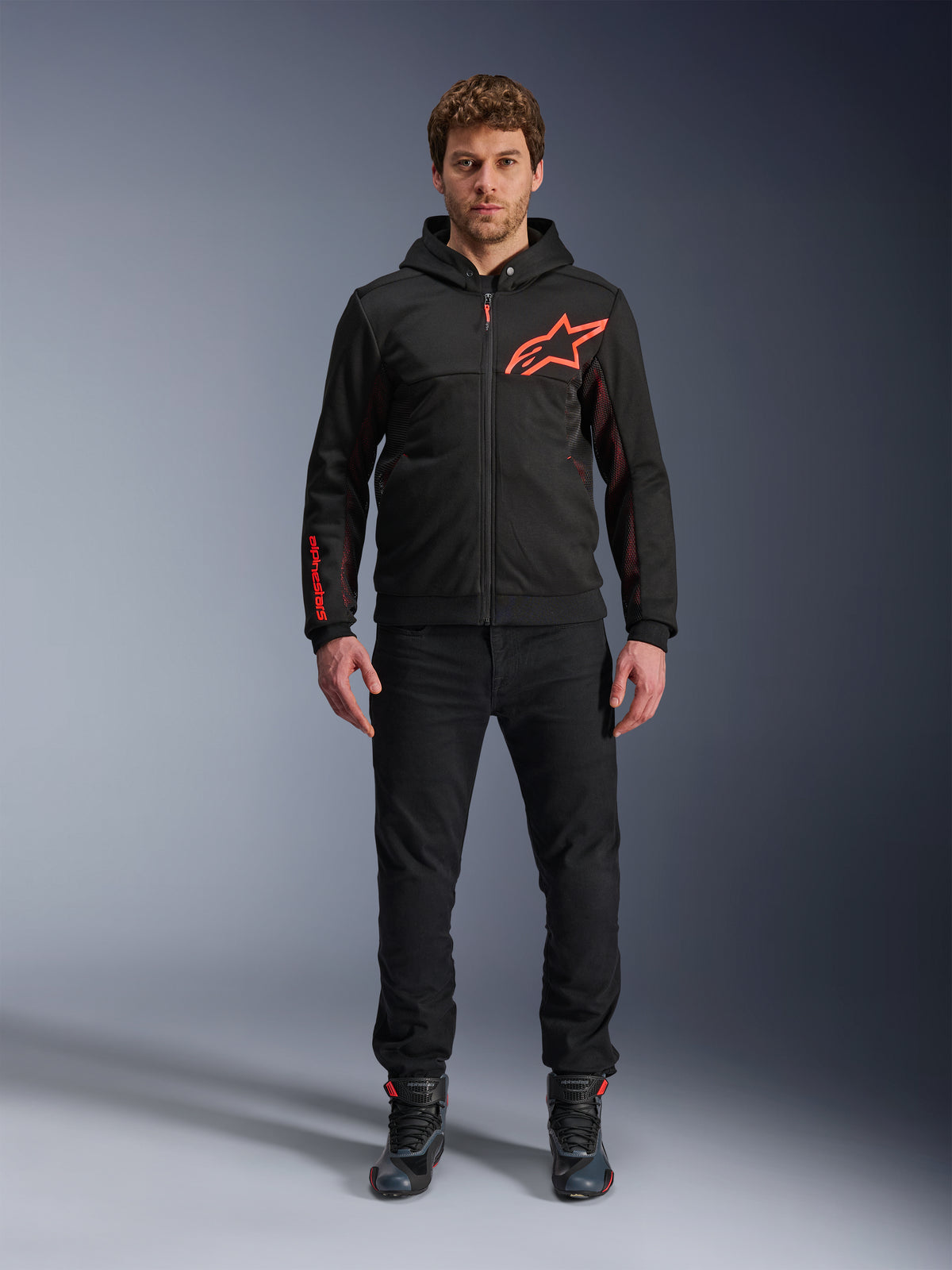 Alpinestars Chrome Air Sport Hoodie, Schwarz und Fluo-Rot, Urbane Motorradjacke mit Tech-Fleece-Konstruktion, mit großem rotem Astars-Logo auf der Brust und Schriftzug am Ärmel, getragen von einem Modell mit schwarzer Hose und Motorradschuhen