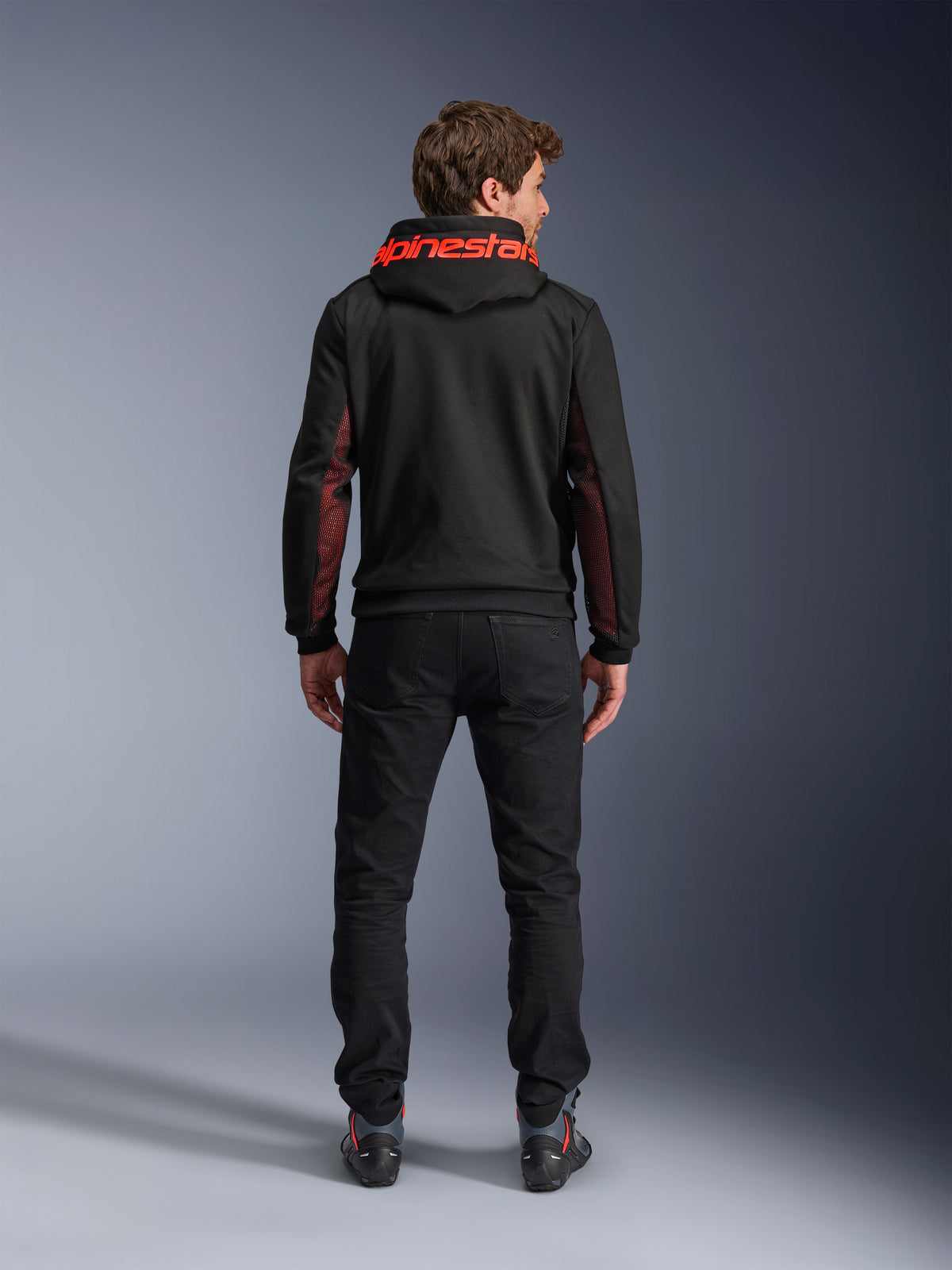 Alpinestars Chrome Air Sport Hoodie, Schwarz und Fluorot, Rückansicht eines Mannes, der eine lässige Motorradjacke trägt, mit rotem Alpinestars-Branding auf der Kapuze und roten Mesh-Einsätzen an den Ärmeln, kombiniert mit einer schwarzen Hose.