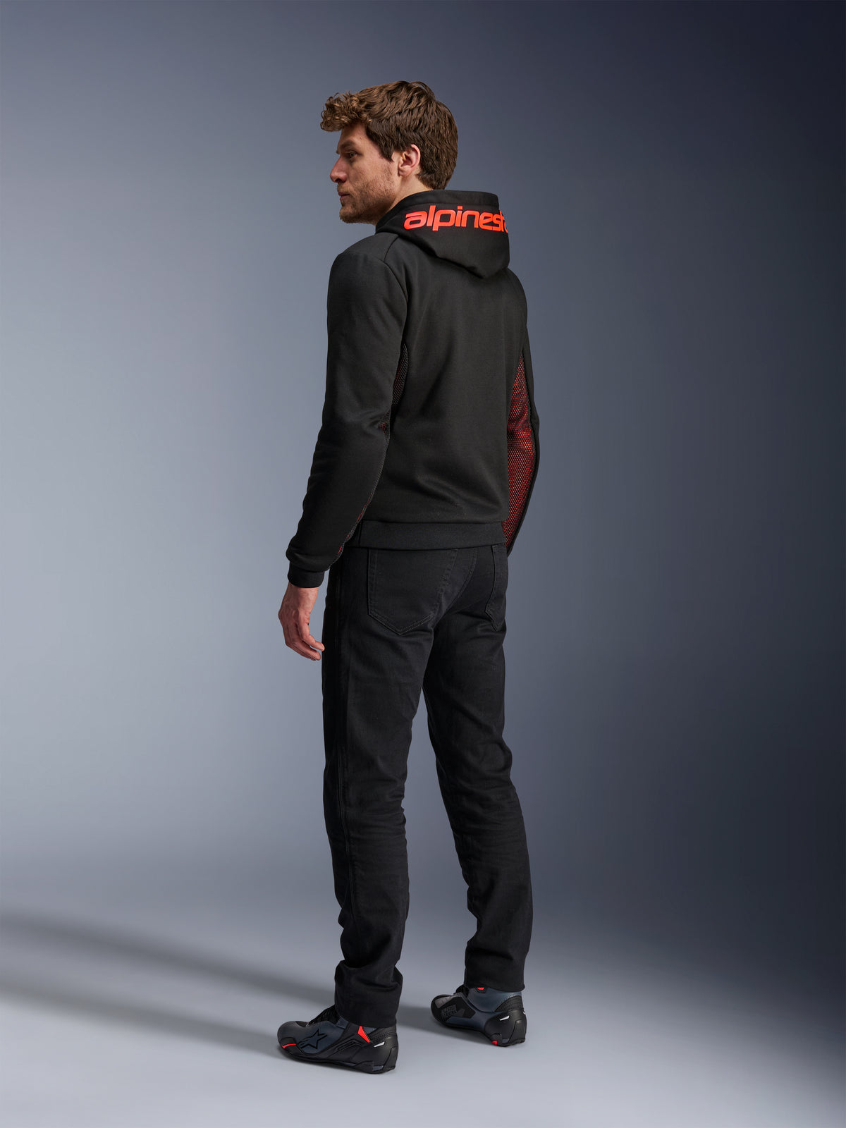 Alpinestars Chrome Air Sport Hoodie, urbaner Motorrad-Hoodie, Schwarz und Fluo-Rot, Rückansicht mit rotem Alpinestars-Branding auf der Kapuze und Mesh-Seiteneinsätzen, getragen mit schwarzer Motorradhose und technischem Schuhwerk