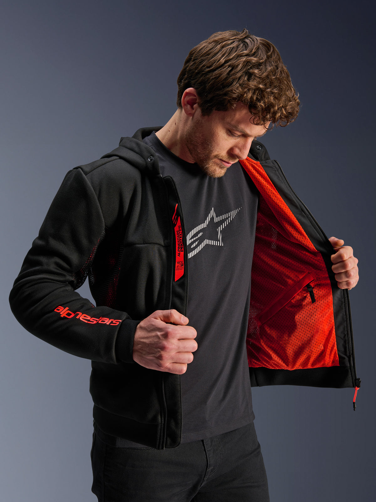 Alpinestars Chrome Air Sport Hoodie in Schwarz und Rot Fluo, männliches Model öffnet die Jacke zur Präsentation des leuchtend roten Mesh-Futters, Alpinestars-Branding am rechten Ärmel und Brustreißverschluss, lässige urbane Motorradjacke für optimale Belüftung