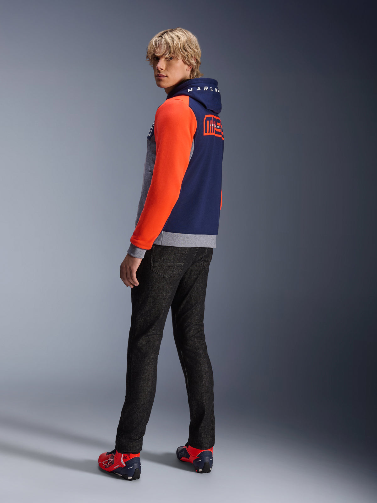 Alpinestars MM93 Chrome V2 Sport Hoodie, Blue Navy Gray Mel Red Fluo, model wearing hoodie with bright red sleeves and MM93 Marc Marquez branding on the back, styled with dark jeans and red riding shoes, casual urban motorcycle gear
