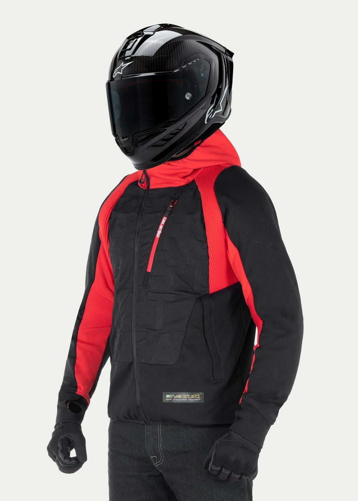 Alpinestars MO.ST.EQ Hybrid Kapuzenjacke, Urbaner Motorrad-Hoodie, Schwarz und Rot, mit schrägem Frontreißverschluss und gerippten Schultereinsätzen, kombiniert mit einem schwarzen Integralhelm und schwarzen Handschuhen, zeitgenössischer Streetwear-Stil