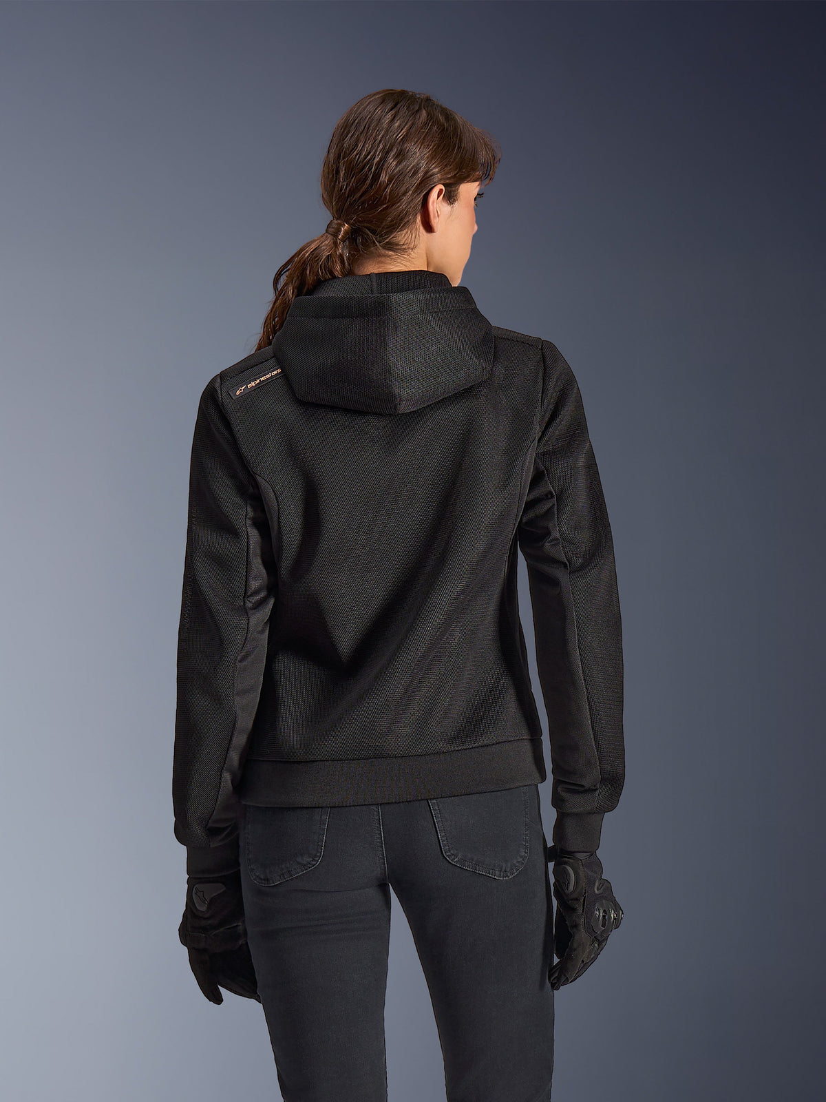 Alpinestars Stella Chrome Superairflow Hoodie, Rückansicht einer Frau, die einen schwarzen technischen 3D-Mesh-Motorrad-Hoodie trägt, mit speziell entwickelter Kapuze und kupferfarbenem Logo auf der Schulter, kombiniert mit schwarzen Motorradhandschuhen und einer schwarzen Hose.