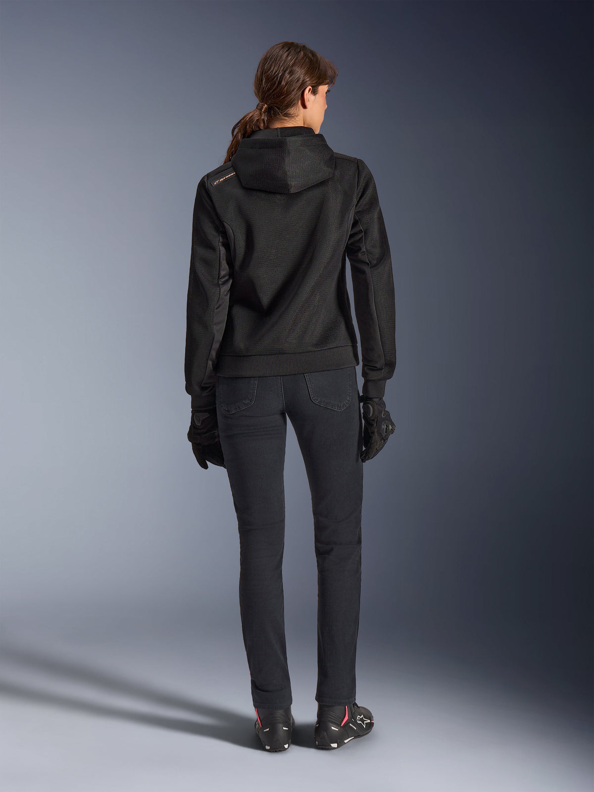 Alpinestars Stella Chrome Superairflow Hoodie, technischer Motorrad-Kapuzenpullover für Damen, schwarzes 3D-Mesh mit kleinem orangefarbenem Logo auf der Schulter, Rückansicht, kombiniert mit schwarzer Motorradhose, schwarzen Handschuhen und Stiefeln, entwickelt für urbanes sportliches Fahren