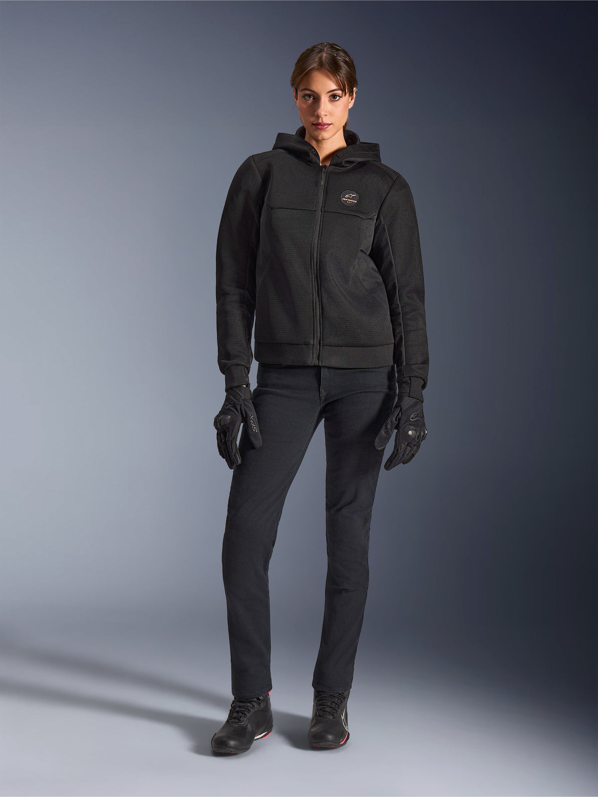Weibliches Modell im Alpinestars Stella Chrome Superairflow Hoodie in Schwarz, kombiniert mit schwarzer technischer Motorradhose und Handschuhen, 3D-Mesh-Textilkonstruktion und kupferfarbenes Logo auf der Brust, urbane Sport-Motorradbekleidung für Damen