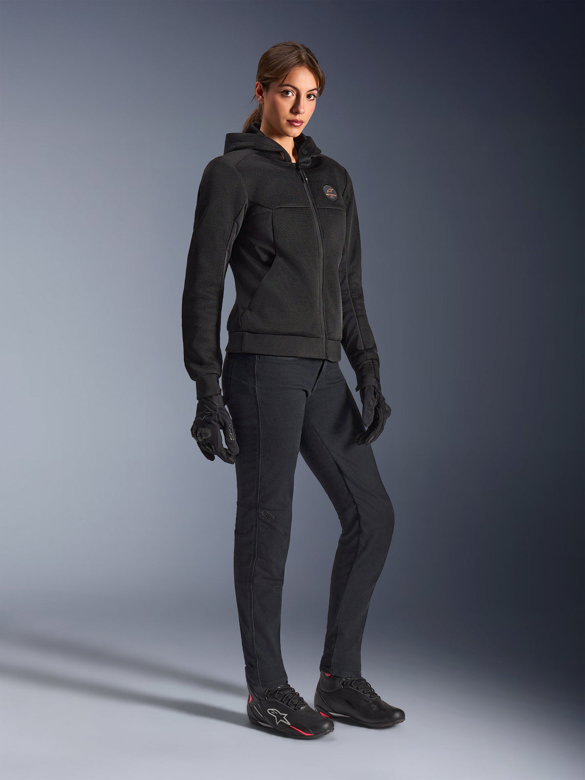 Motorradfahrerin im Alpinestars Stella Chrome Superairflow Hoodie in Schwarz, technischer Motorrad-Hoodie mit 3D-Mesh-Konstruktion, kombiniert mit schwarzer Motorradhose, Handschuhen und sportlichen Motorradschuhen, urbane Sport-Ästhetik.