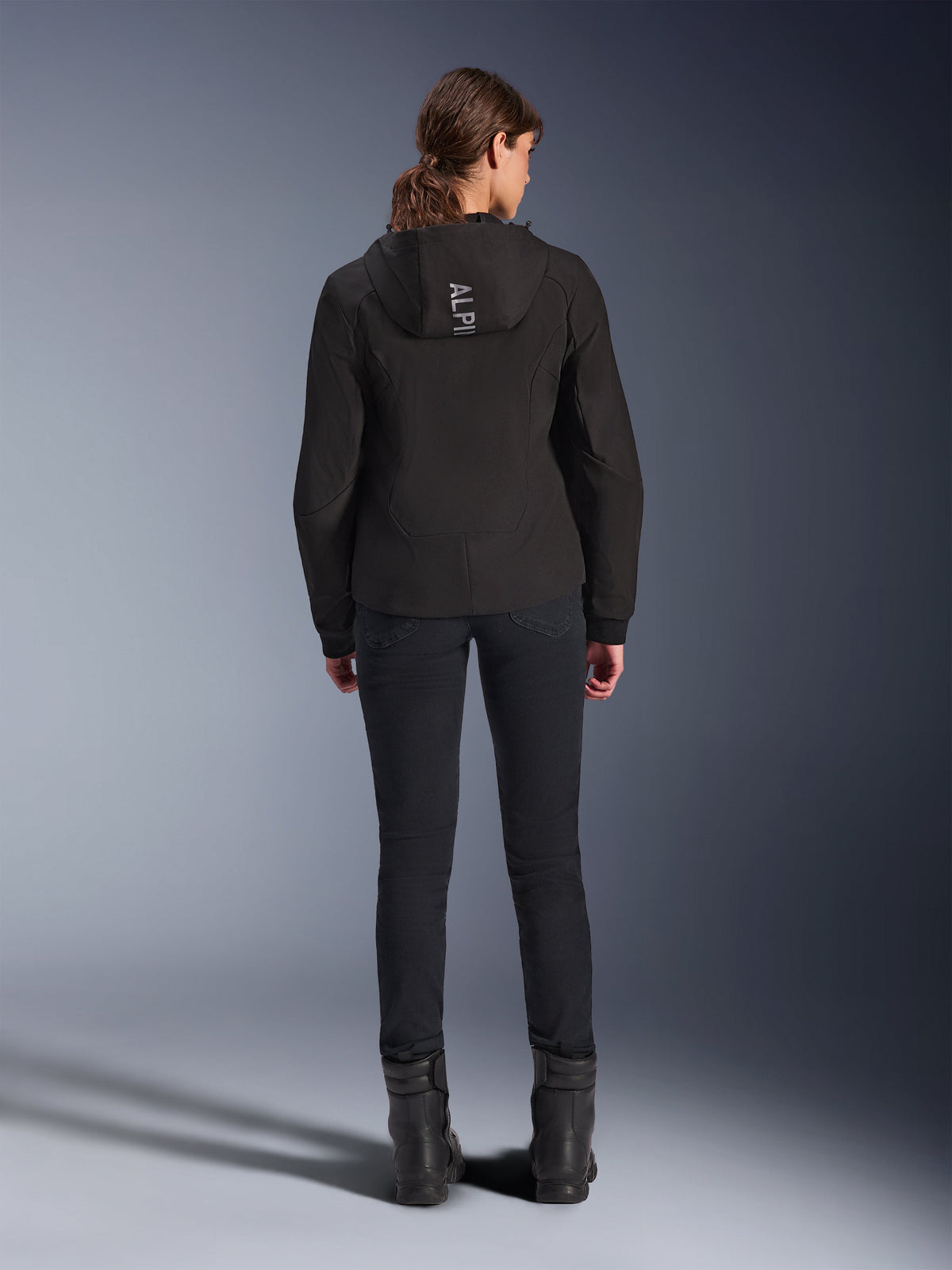Alpinestars Stella Radium Tech Hoodie für Damen, Schwarz, Rückansicht eines Models, das einen leichten, 3-lagigen, winddichten Tech-Hoodie mit Alpinestars-Logo auf der Kapuze trägt, kombiniert mit schwarzer Hose und Stiefeln, urbane Motorradbekleidung