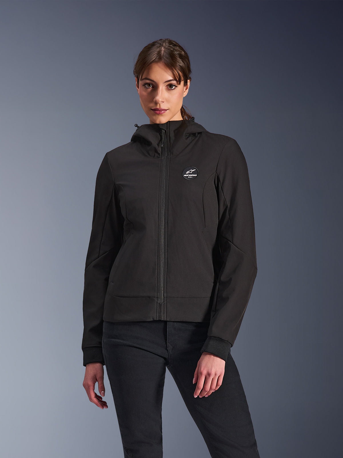 Alpinestars Stella Radium Tech Hoodie, Damen-Motorrad-Kapuzenjacke, Schwarz, Reißverschluss-Design mit rundem Brustlogo und Kapuze, technische urbane Fahrerausrüstung, von einem Model getragen