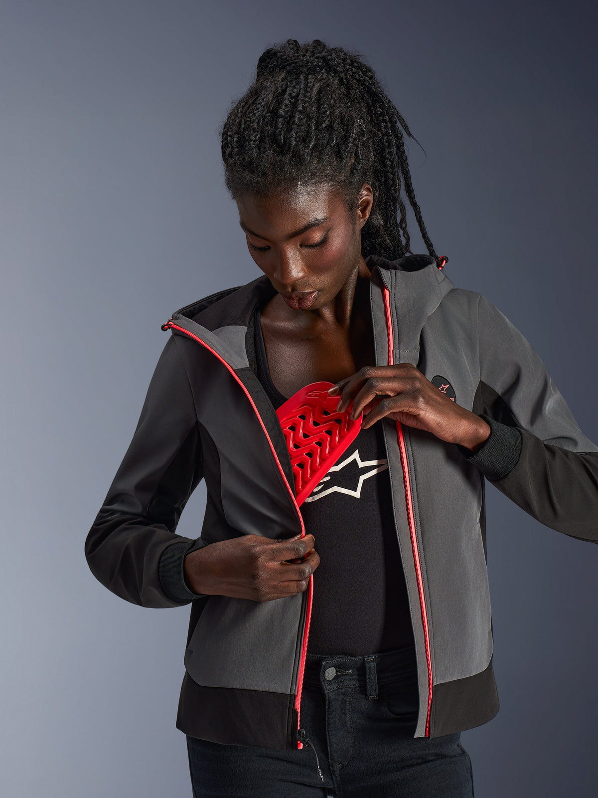 Alpinestars Stella Radium Tech Hoodie für Damen in Tiefgrau und Schwarz, urbaner Motorrad-Hoodie mit pinken Reißverschluss-Akzenten, Modell demonstriert den herausnehmbaren roten Nucleon Flex Plus Protektor.