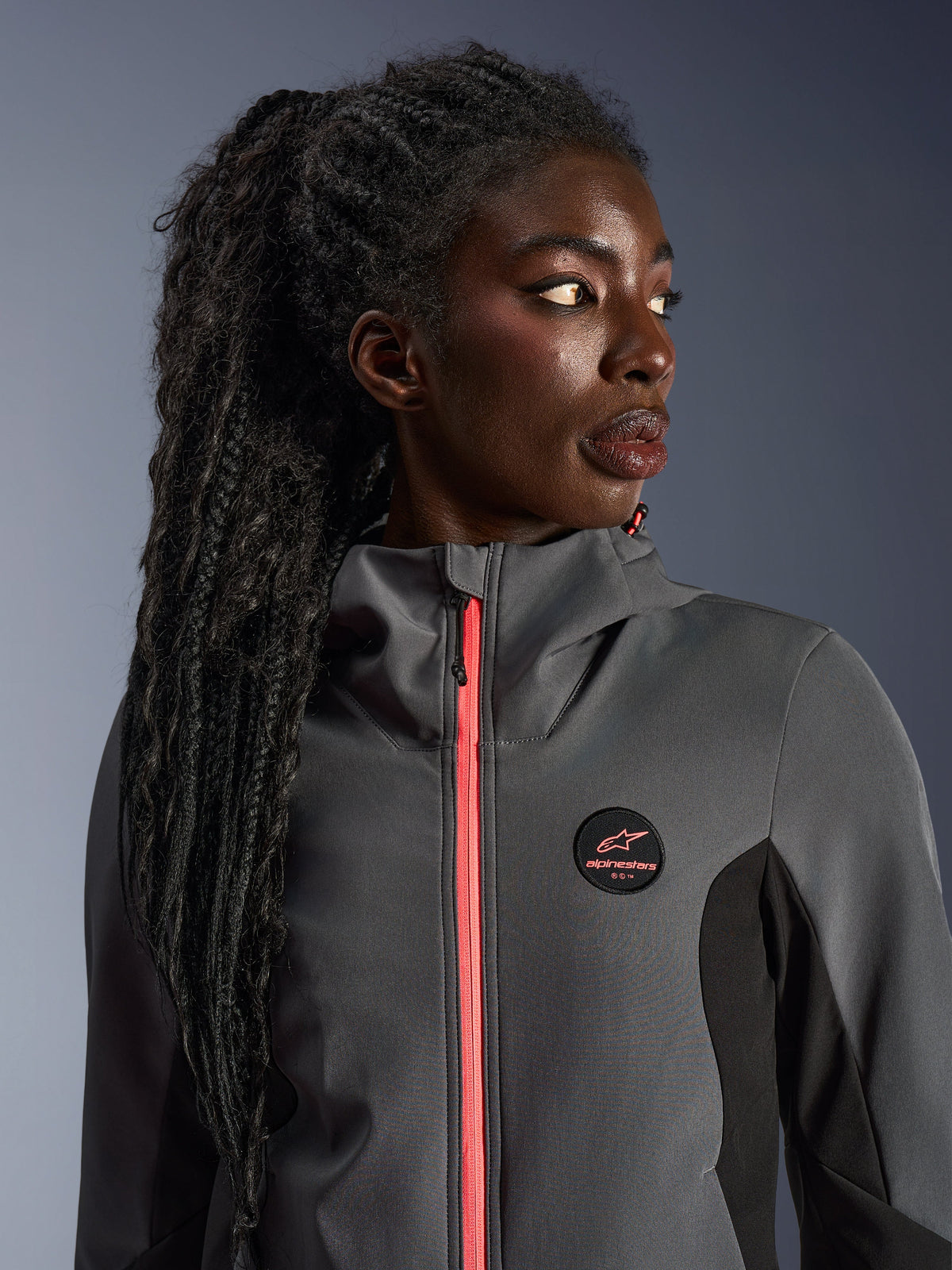 Weibliches Model im Alpinestars Stella Radium Tech Hoodie in Dunkelgrau und Schwarz, mit leuchtend korallenfarbenem Reißverschluss, rundem Brust-Logo und winddichter High-Neck-Shell, lässige urbane Motorradausrüstung für Damen