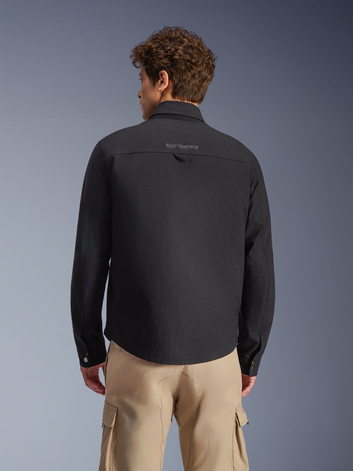 Alpinestars Bryon Water Resistant Overshirt, Schwarz, Rückansicht am Model, mit gesticktem Logo auf der Rückenpasse und praktischer Aufhängeschlaufe, kombiniert mit sandfarbenen Cargohosen, urbane Motorradbekleidung mit klassischer Workwear-Ästhetik