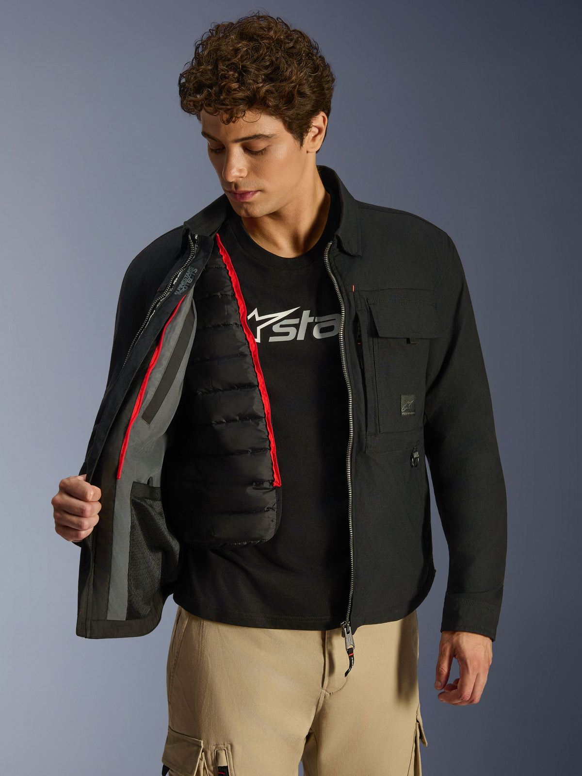 Alpinestars Bryon WR Overshirt, Schwarz, urbane Motorradjacke, offen getragen, zeigt eine herausnehmbare schwarze gesteppte Thermoweste mit rotem Rand, kombiniert mit beiger Cargohose und einem Logo-T-Shirt, funktionales, von Workwear inspiriertes Design