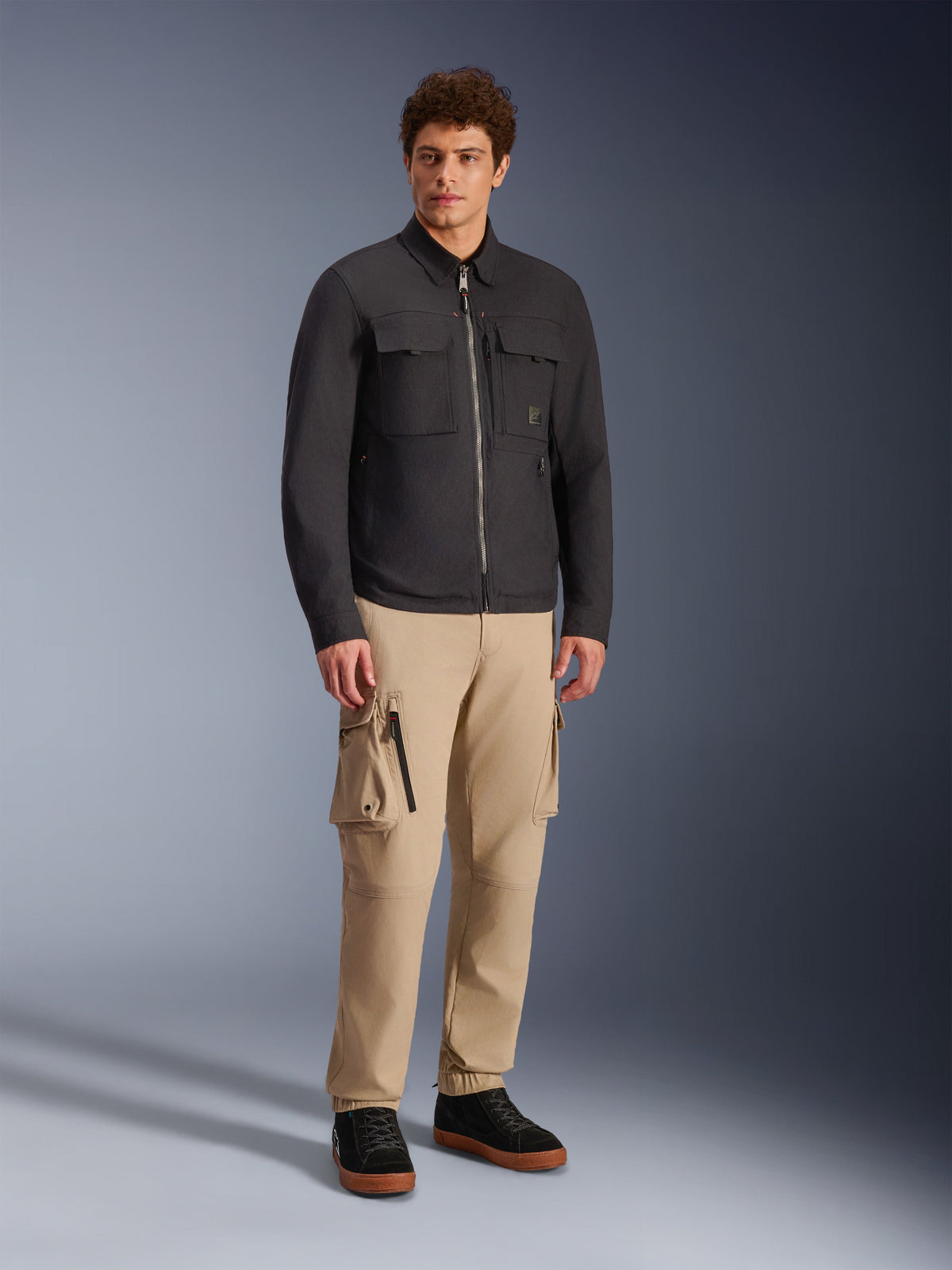 Alpinestars Bryon Water Resistant Overshirt, Schwarz, getragen von männlichem Model mit khakifarbener Cargohose und schwarzen Schuhen, urbane Motorradjacke im Workwear-inspirierten Design, Ganzkörper-Studioaufnahme