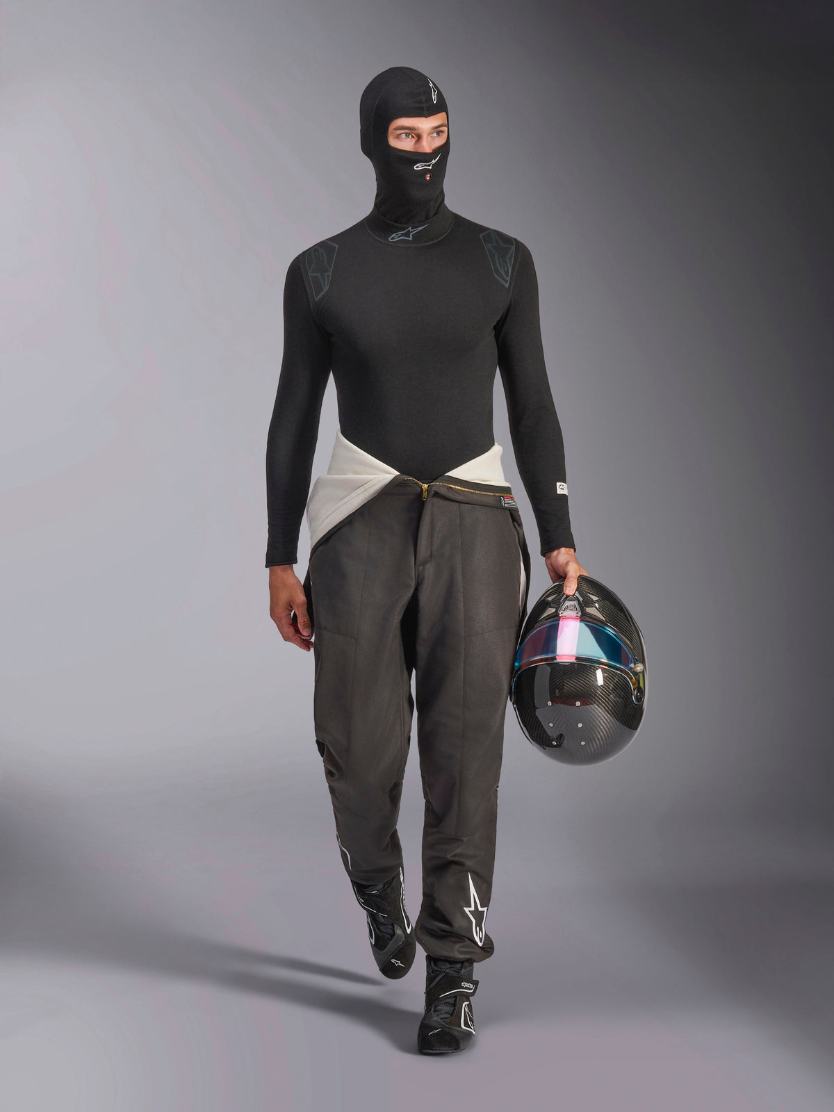Alpinestars ZX Evo V3 Sturmhaube, Schwarz, getragen von einem Model in einem kompletten Rennsport-Base-Layer-Outfit mit passendem langärmeligem Oberteil und grauer Rennhose, hält einen Carbon-Helm mit schillerndem Visier, professionelle Automobil-Rennausrüstung