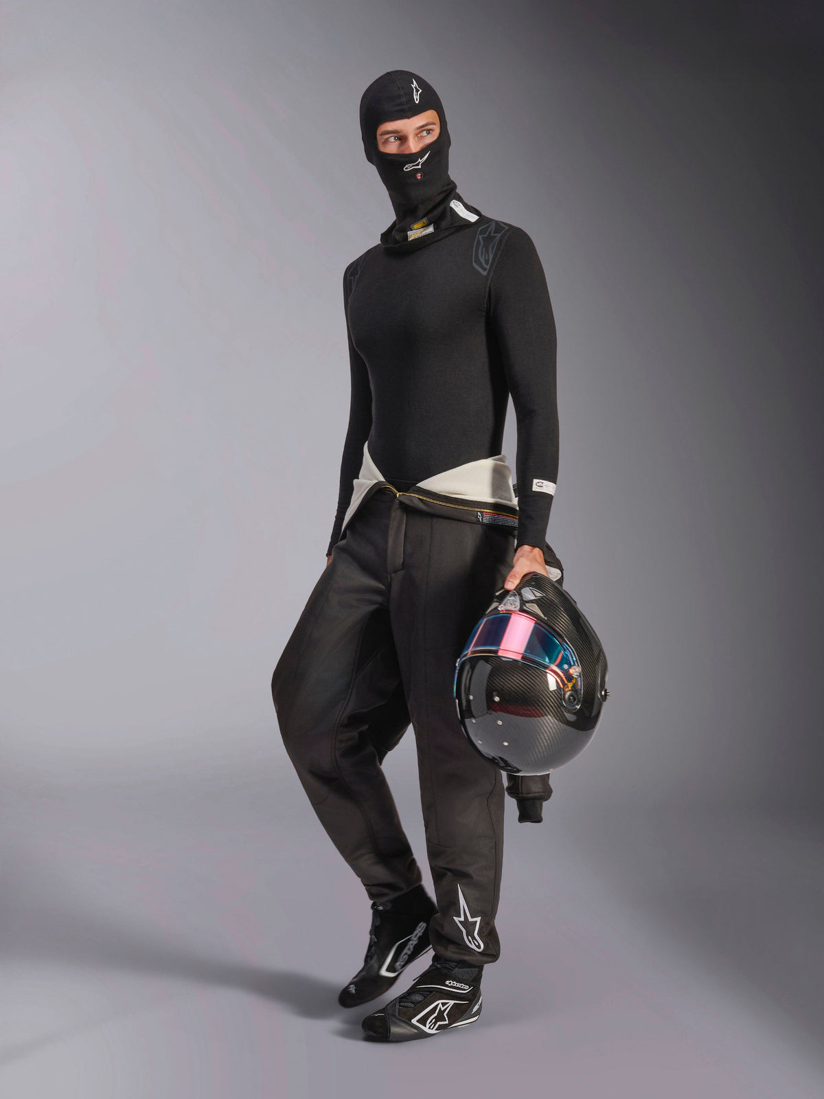 Alpinestars ZX Evo V3 Sturmhaube, Schwarz, getragen von einem Fahrer mit kompletter Auto-Rennsport-Unterbekleidung, mit weißem Logo auf der Stirn, hält einen Carbon-Helm mit schillerndem Visier, professionelle feuerfeste Motorsportbekleidung.