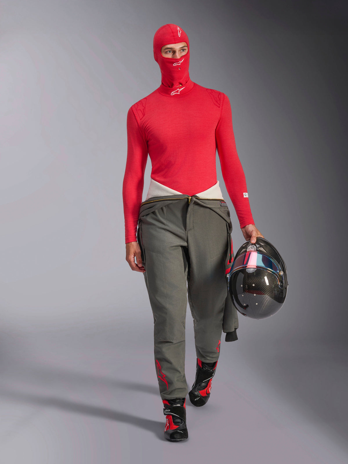 Race driver wearing Alpinestars ZX Evo V3 Balaclava in Red, matching red long-sleeve base layer, grey racing pants, black and red racing boots, carrying a carbon fiber helmet, professional auto racing gear set.