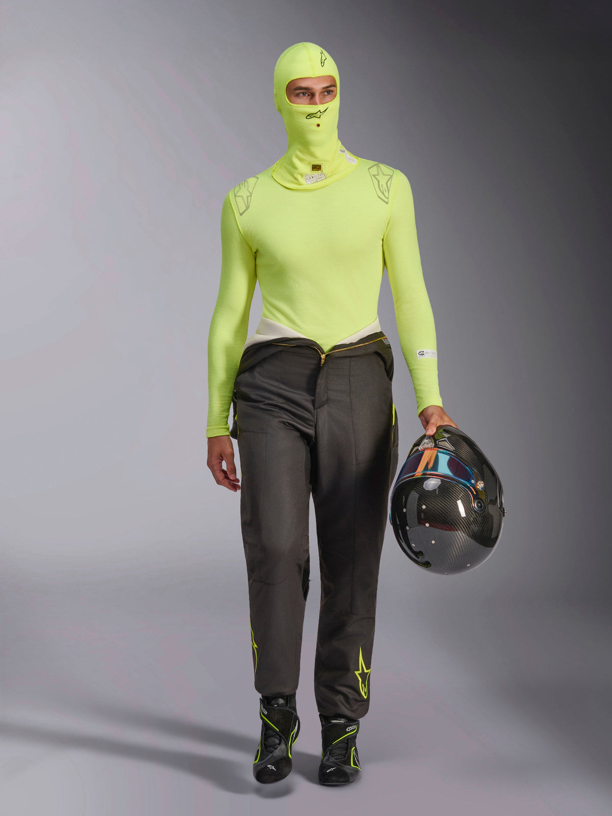 Alpinestars ZX Evo V3 Sturmhaube in Yellow Fluo, kombiniert mit passendem langärmeligem Base-Layer-Oberteil und dunkelgrauer Rennhose, männliches Model hält einen Carbon-Helm, professionelle Autorennsport-Ausrüstung