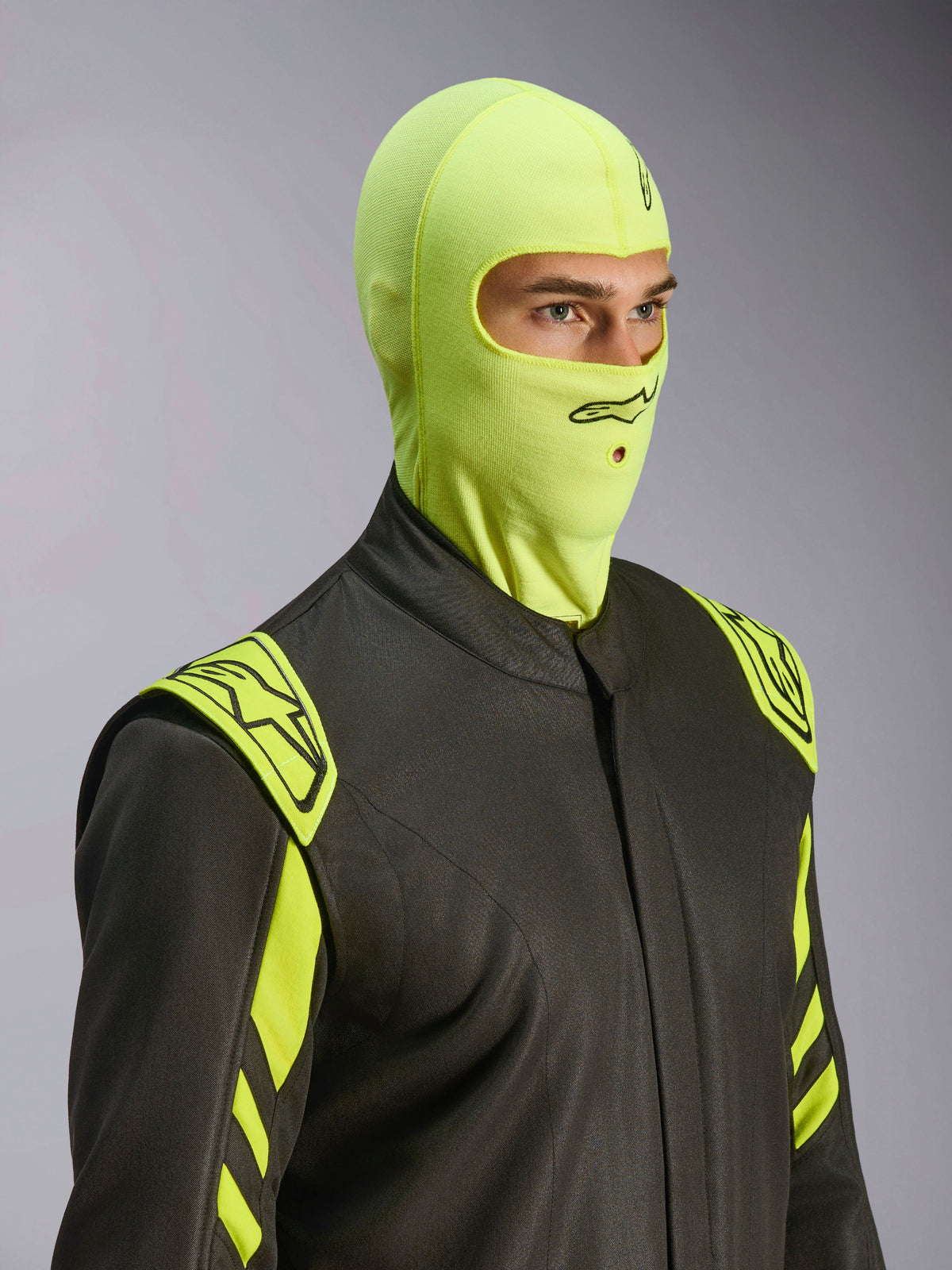 Männliches Model in der Alpinestars ZX Evo V3 Sturmhaube in Yellow Fluo, flammenhemmende Rennsport-Kopfhaube, kombiniert mit einem dunkelgrauen Autorennanzug mit neongelben Akzenten, mit schwarzem Alpinestars-Logo und Mundöffnung für Trinkschlauch, professionelle Motorsport-Schutzausrüstung.