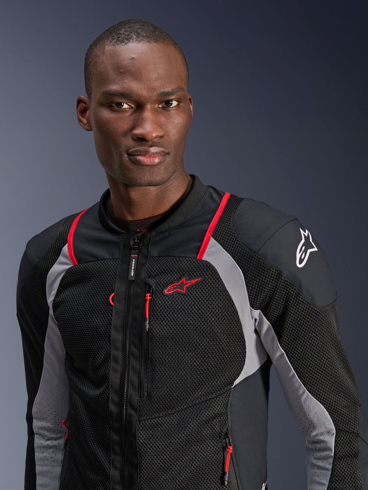 Mann in Alpinestars Troop-Air Pro Jacke, Schwarz, Ash Gray und Bright Red, technische Panzer-Air-Jacke mit atmungsaktiven Mesh-Einsätzen, roten Paspel-Akzenten und Alpinestars-Logo auf der Schulter, moderne Urban-Motorradausrüstung