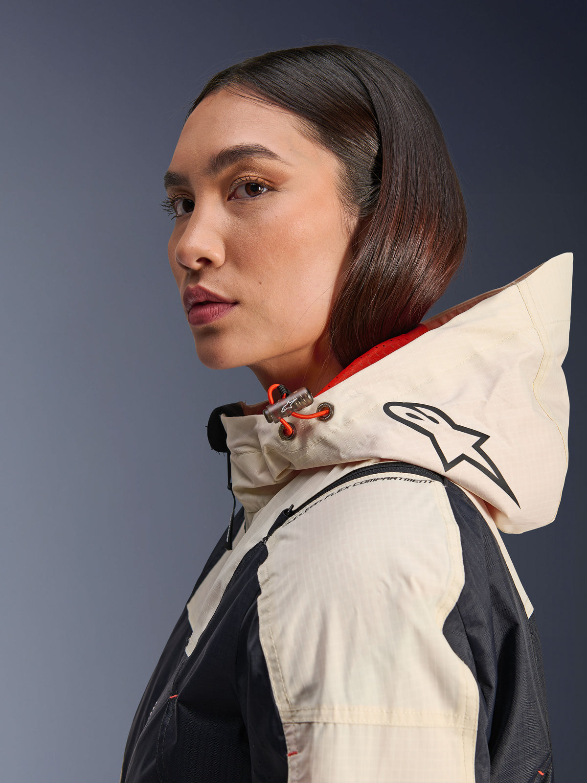 Alpinestars Molly Air Jacke, Black Almond, moderne urbane Motorradjacke, Seitenprofil einer Frau mit Kapuze samt Alpinestars-Logo und orangefarbenen Kordelzug-Akzenten, Ripstop-Gewebe-Textur