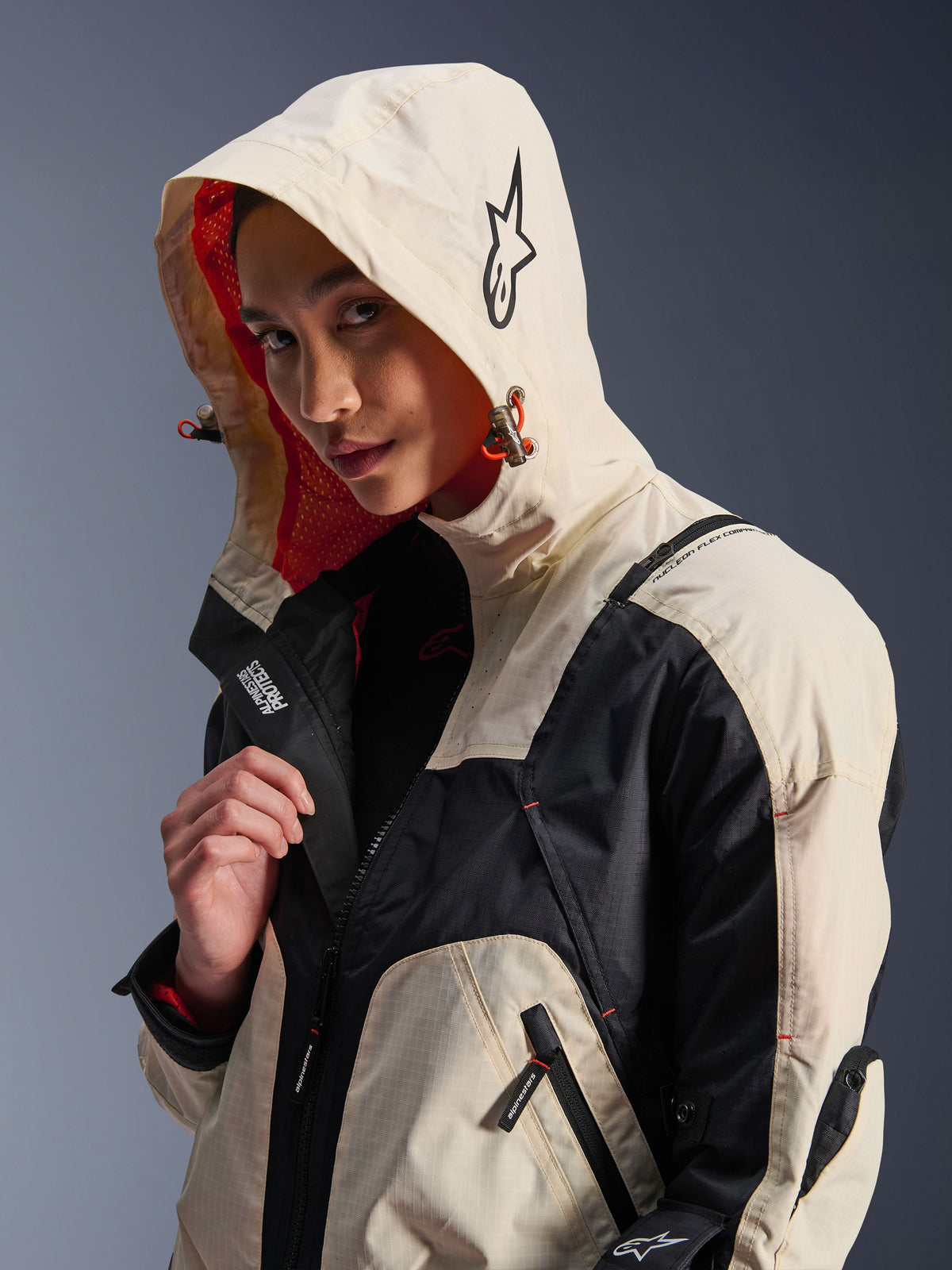 Alpinestars Molly Air Jacke in Black Almond, getragen von einem weiblichen Model mit aufgesetzter Kapuze, mit cremefarbenem und schwarzem Color-Blocking, leuchtend orangefarbenem Mesh-Futter und technischen urbanen Designelementen wie Stern-Logo und Reißverschlussdetails.
