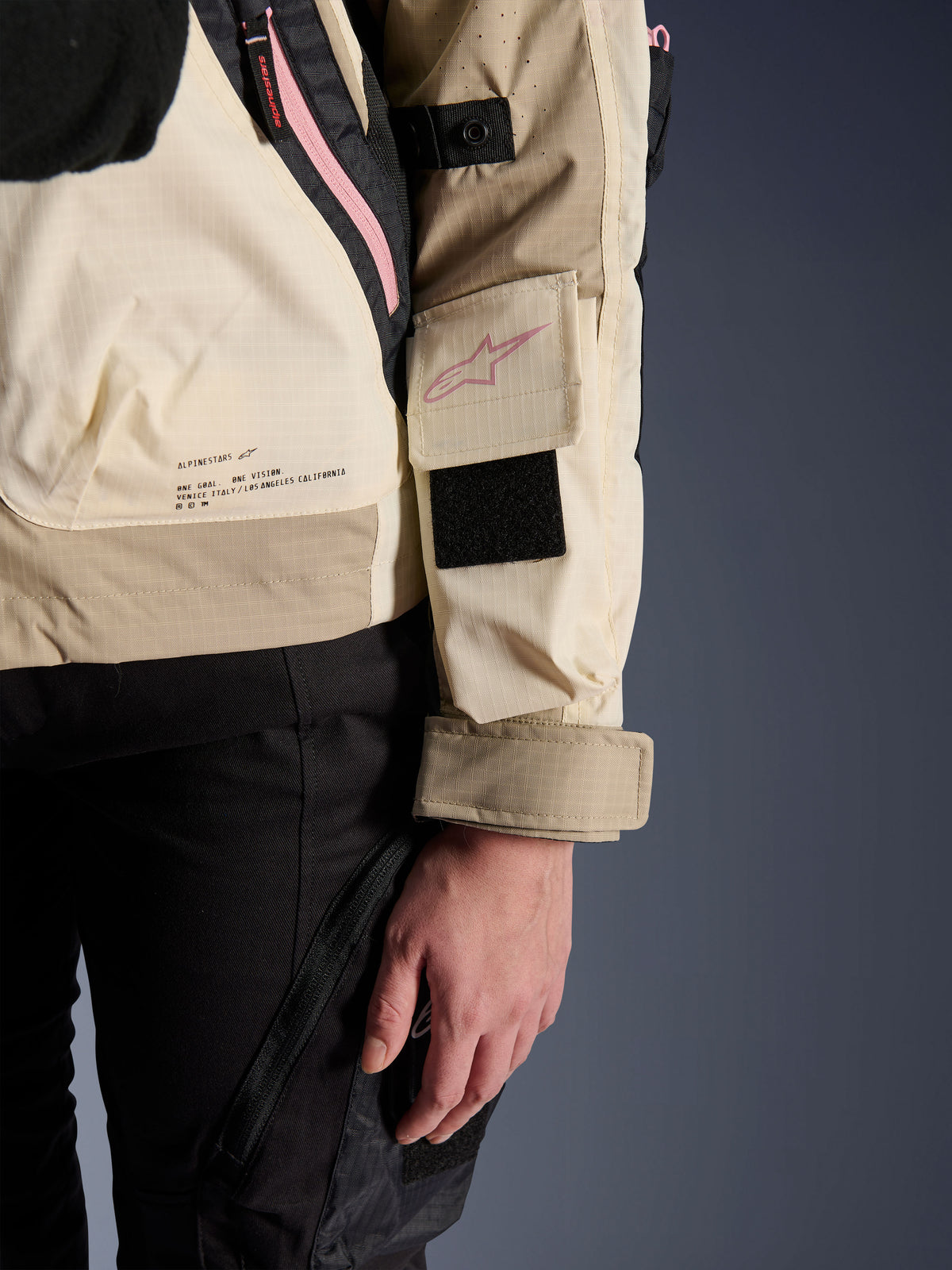 Alpinestars Molly Air Jacke, Urban-Motorradjacke für Damen, Sand Pink Almond, Nahaufnahme der Seitenansicht mit Ärmeltasche und pinkfarbenem Logo, schwarzem Klettverschluss-Patch und Text der Markenmission am unteren Rücken, leichtes Ripstop-Material