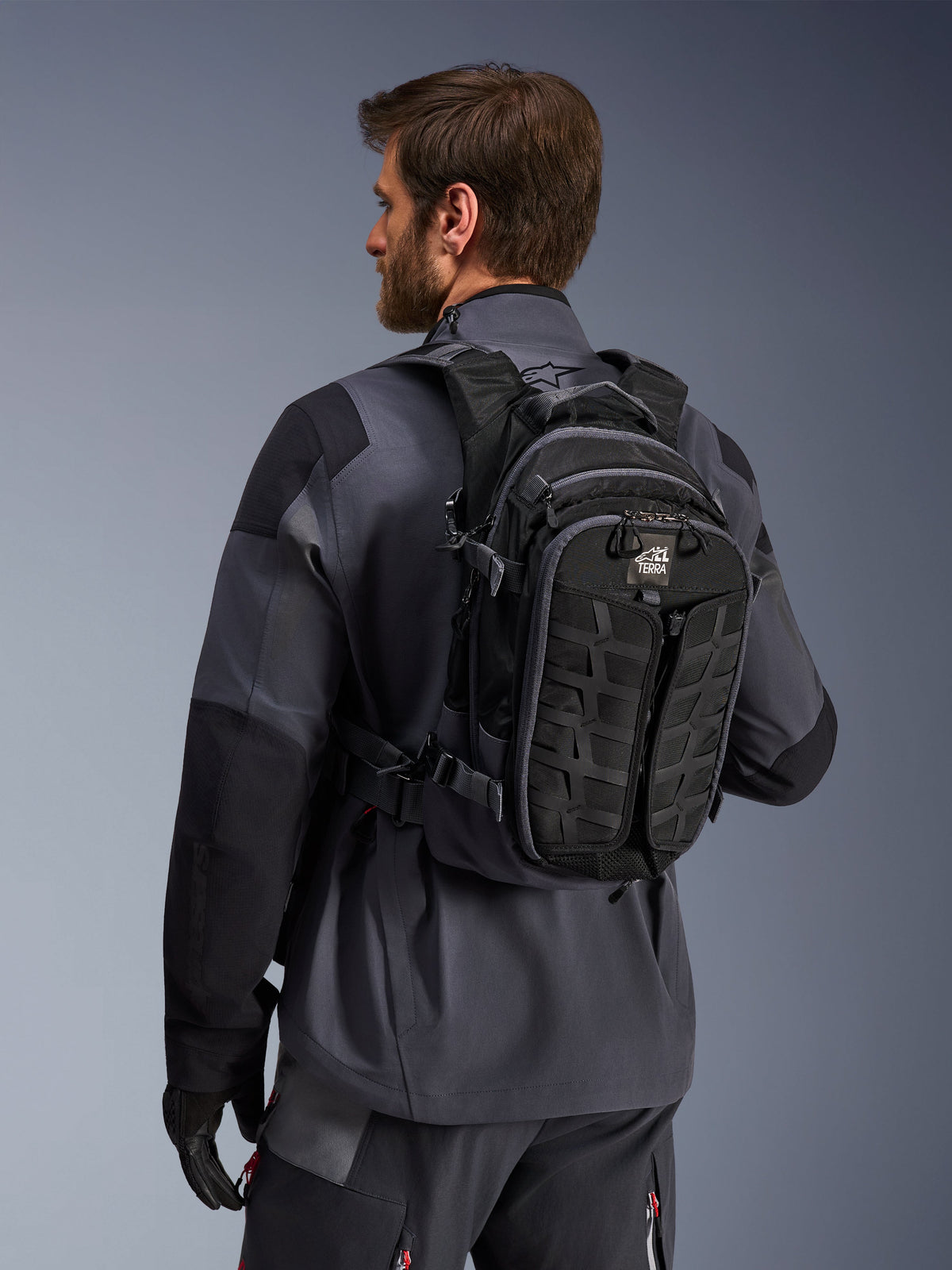 Alpinestars AT-15 Rucksack, Adventure-Motorradrucksack, Schwarz und Dunkelgrau, getragen von einem Fahrer in einer grauen Jacke, mit technischem Design, MOLLE-Gurtband und „TERRA“-Branding, ergonomische Offroad-Ausrüstung