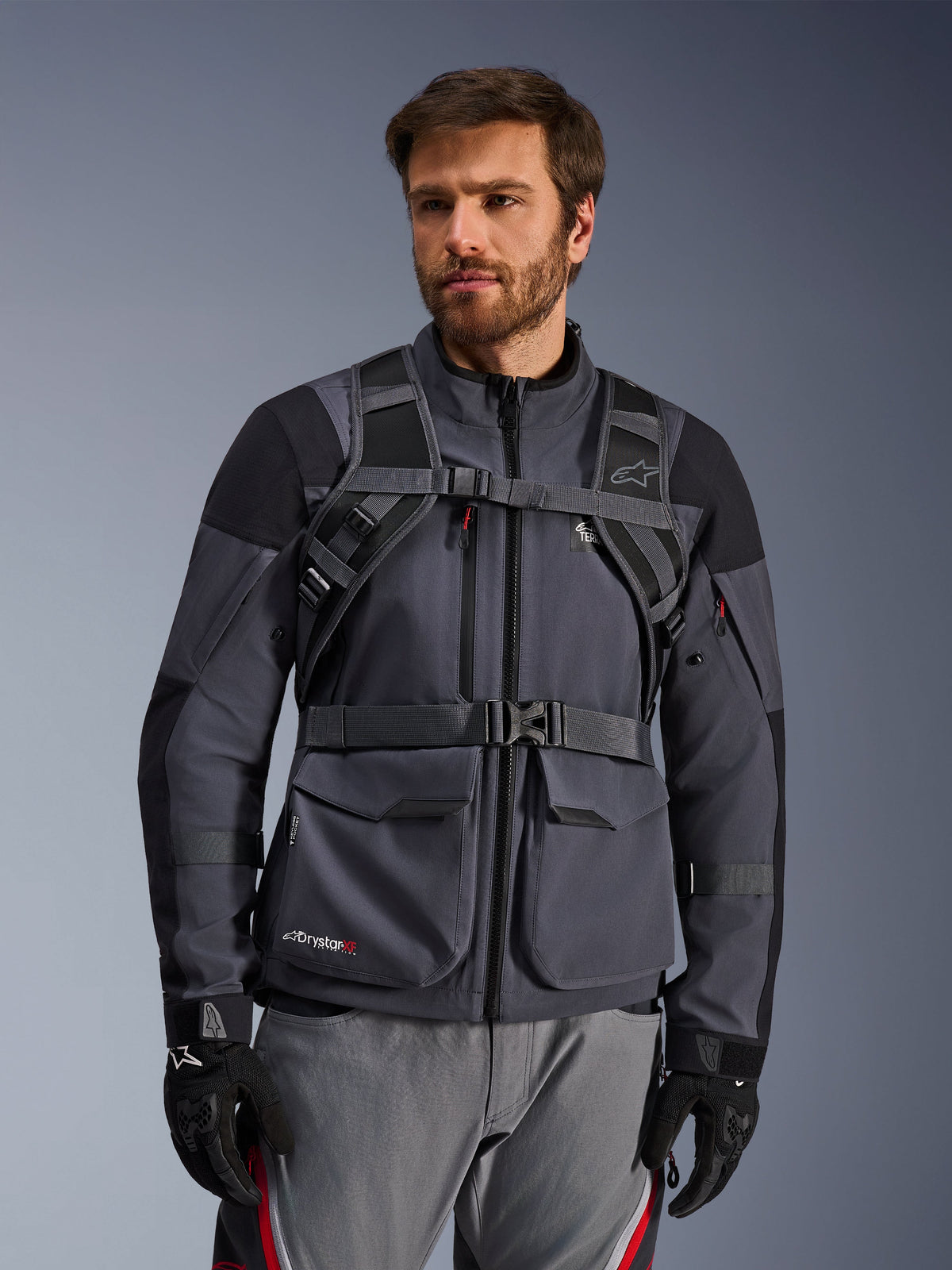 Alpinestars AT-15 Rucksack-Gurtsystem, Schwarz Dunkelgrau, Vorderansicht getragen von einem Fahrer über einer grauen Motorradjacke, mit magnetischem Brustverschluss und sicherem Hüftgurt, kombiniert mit schwarzen Handschuhen für Adventure-Motorradsport