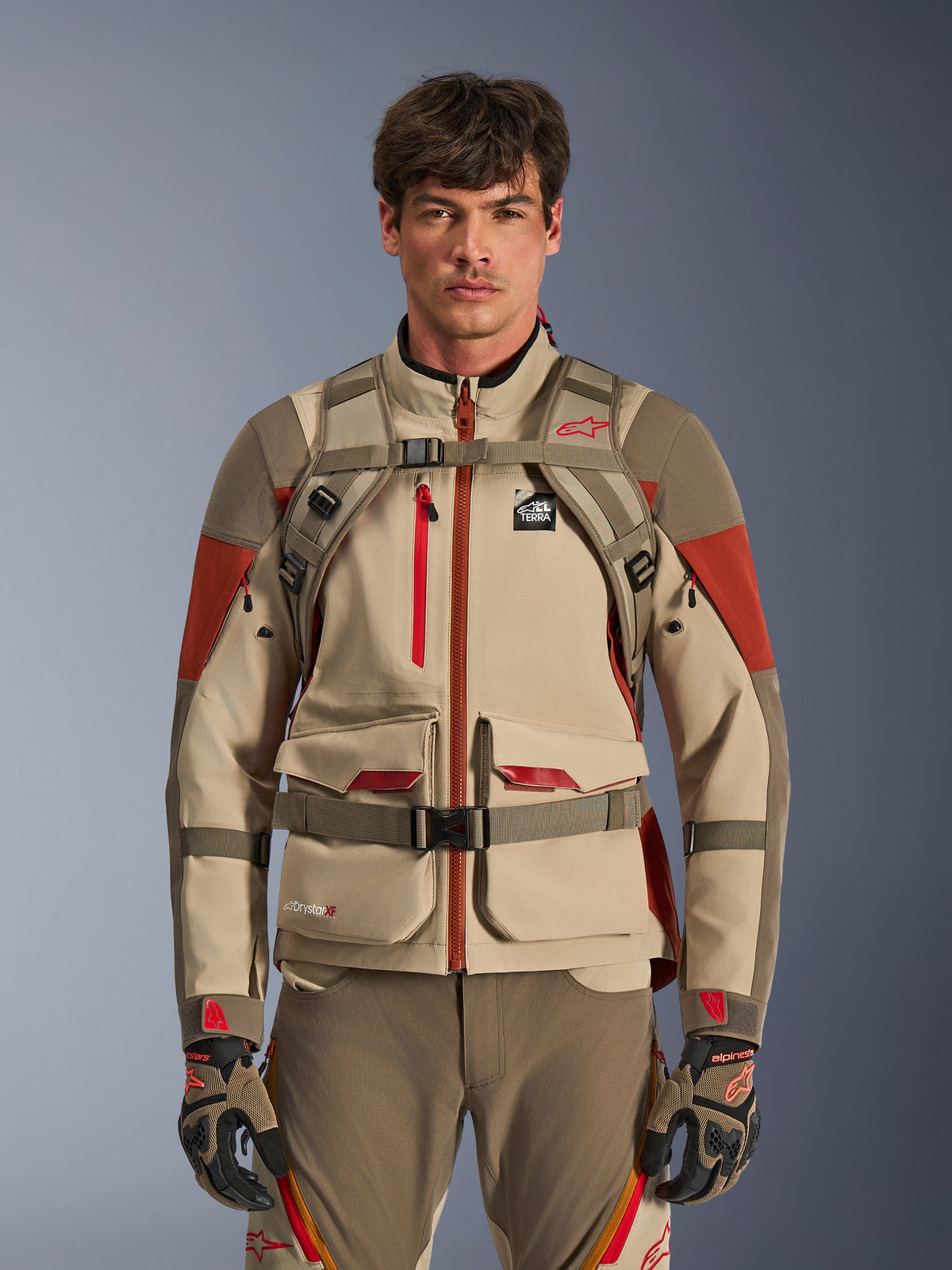 Alpinestars AT-15 Rucksack in Aluminum Walnut, getragen von einem Adventure-Fahrer, Frontansicht mit sichtbaren Brust- und Hüftgurten und Magnetverschluss, kombiniert mit sandfarbener und roter Adventure-Motorradjacke und -hose, technisches Offroad-Ausrüstungsset
