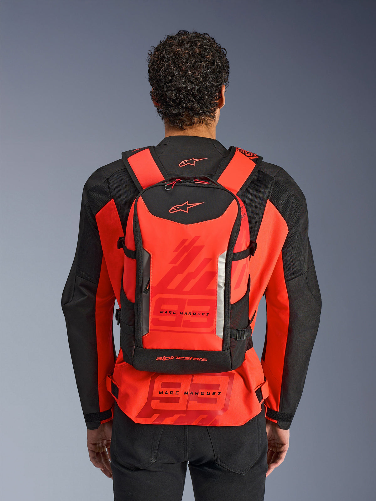 Alpinestars MM93 AMP3 Rucksack, technischer Motorradrucksack, Neonrot und Schwarz, von einem Fahrer von hinten über einer passenden rot-schwarzen MM93 Rennjacke getragen, mit Marc Marquez Branding und Alpinestars-Logos.