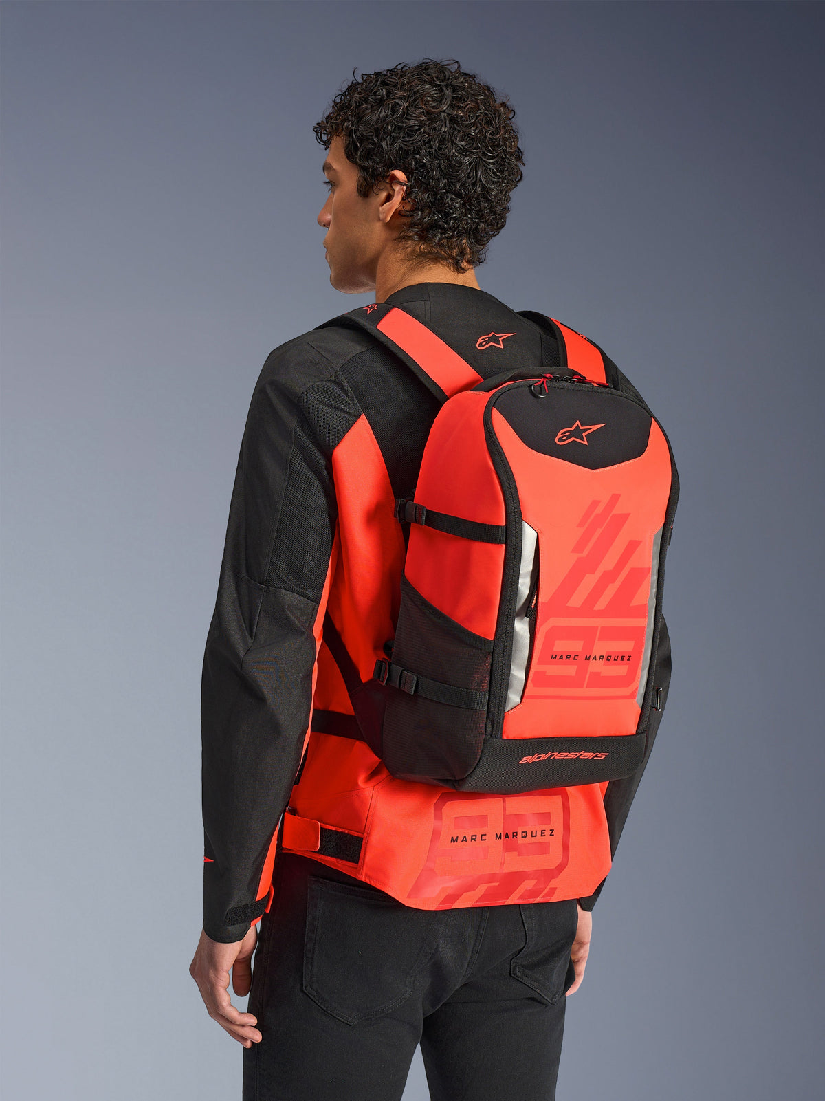 Alpinestars MM93 AMP3 Rucksack, technischer Motorradrucksack, Leuchtrot und Schwarz, von einem Modell von hinten über einer passenden rot-schwarzen Jacke getragen, mit Marc Marquez „93“-Grafiken, verstellbaren Gurten und einem schlanken, sportlich inspirierten Design.