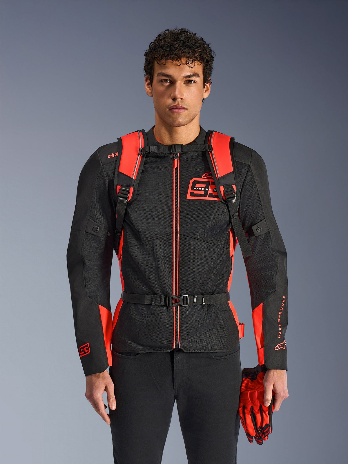 Mann mit Alpinestars MM93 AMP3 Rucksack, Leuchtrot und Schwarz, mit verstellbaren Schultergurten und Brustgurt mit Schnalle, kombiniert mit passender MM93 Motorradjacke und Handschuhen, technische Ausrüstung aus der Marc Marquez Kollektion