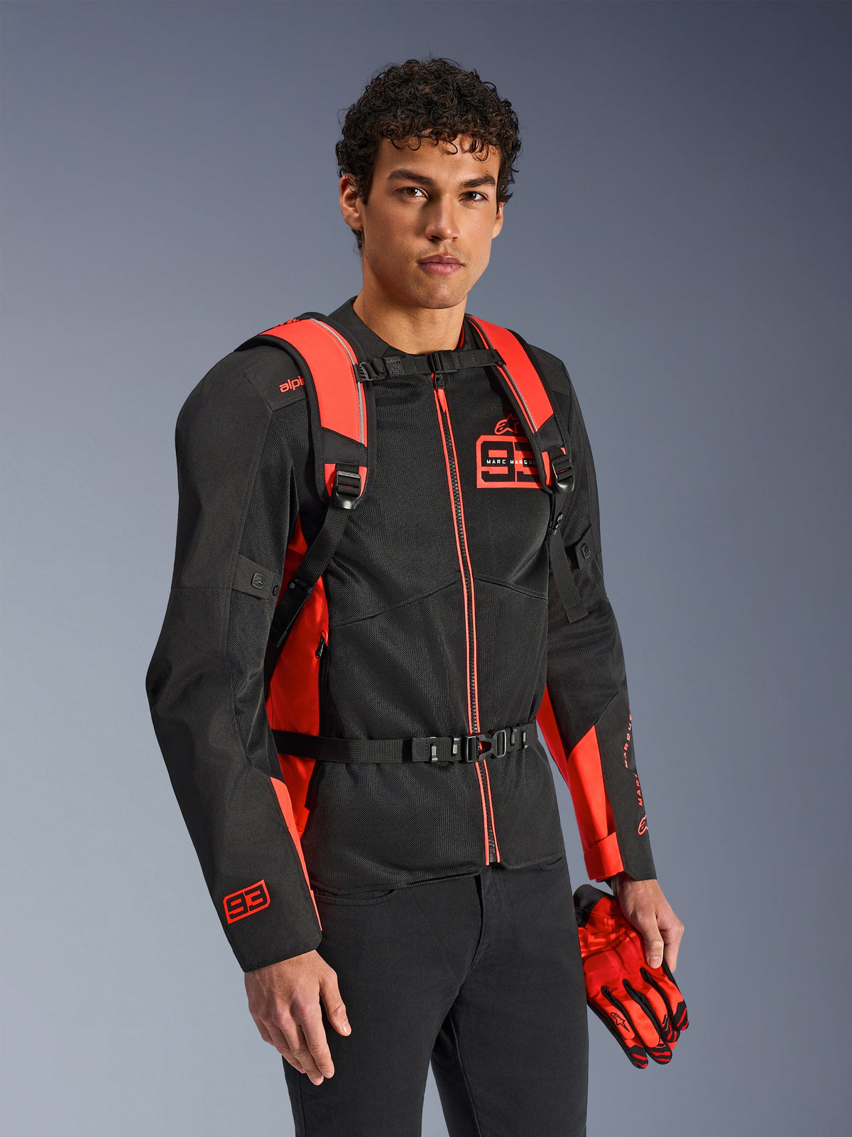 Alpinestars MM93 AMP3 Rucksack, Rot Fluo und Schwarz, getragen von einem Model mit passender schwarz-roter MM93 Motorradjacke und Handschuhen, Vorderansicht mit Fokus auf die verstellbaren Schultergurte und die Brustschnalle, technische vom Rennsport inspirierte Ausrüstung.