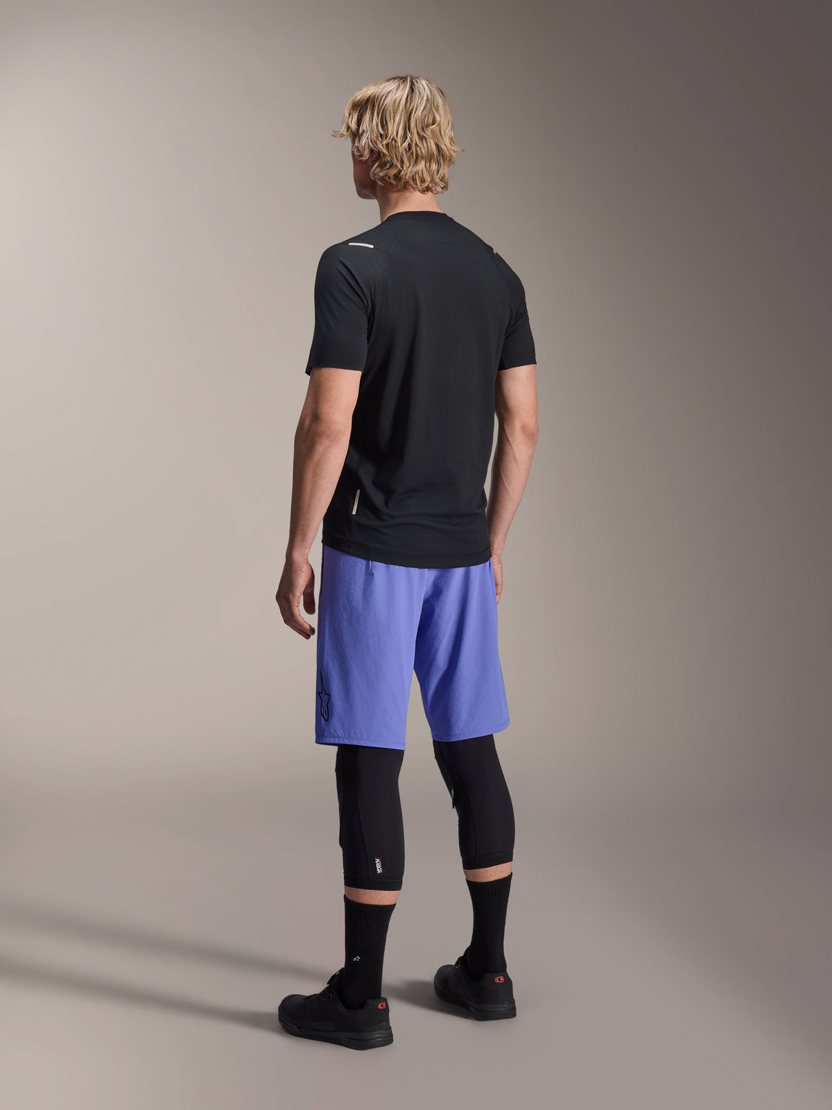 Male model from behind wearing Alpinestars A-Aria Lite Shorts in Baja Blue, paired with a black short-sleeve jersey and black knee protectors, professional MTB cycling apparel for trail riding.