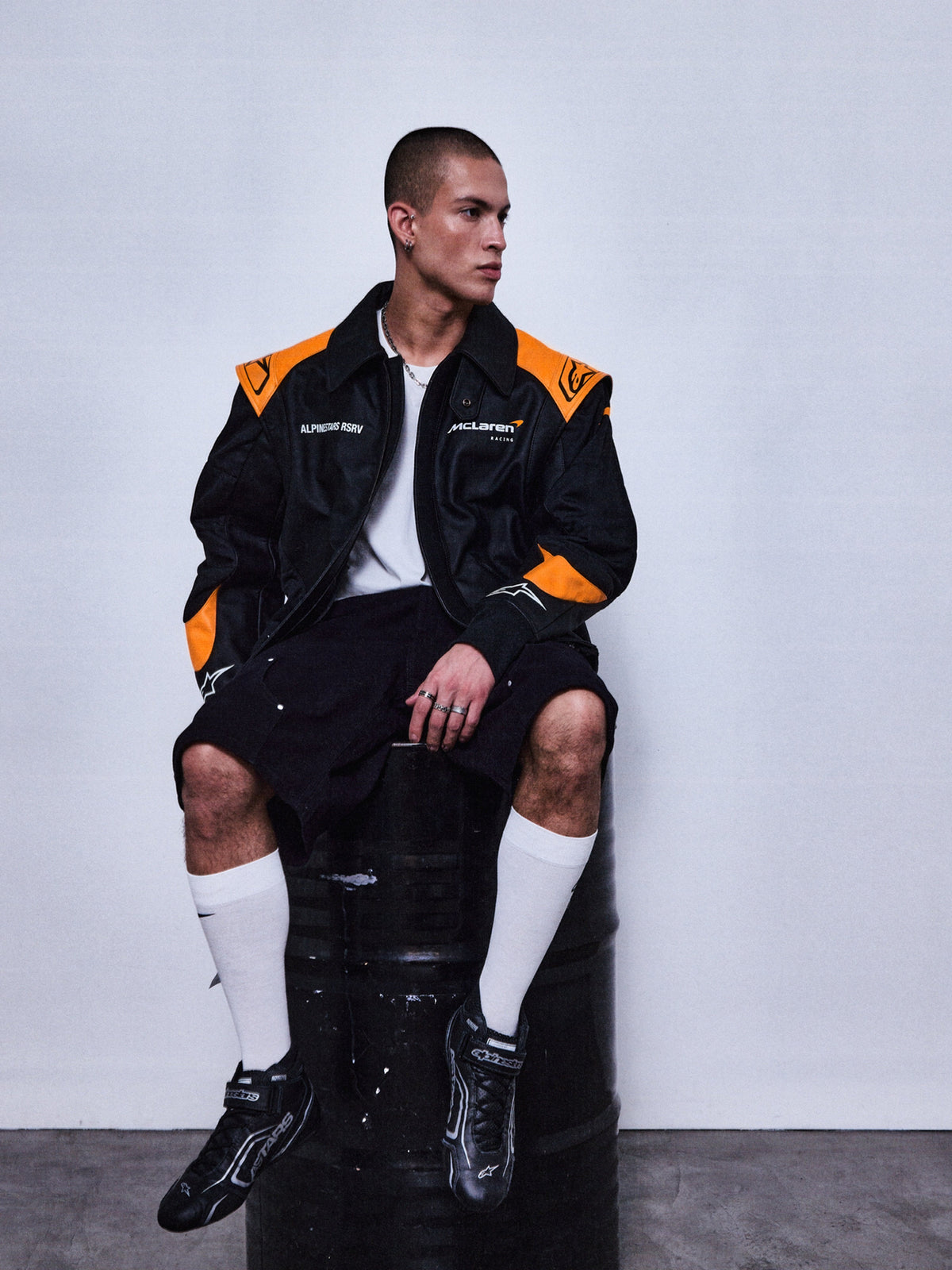 Alpinestars RSRV Track Star Lederjacke, Schwarz und Orange, Premium-Motorradjacke, Person trägt sportliche Jacke mit Logos, urbaner Style, schwarze Shorts und weiße Socken