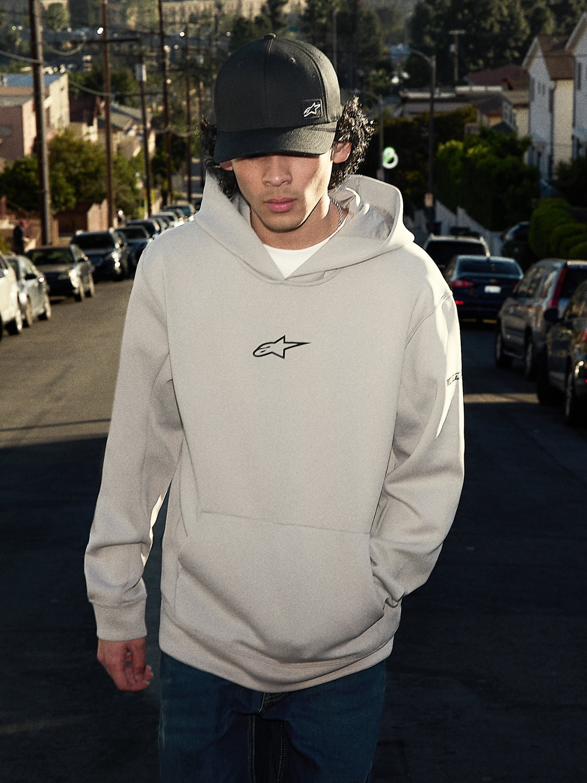 Alpinestars Insight Hoodie, lässige Lifestyle-Streetwear, Cement-Grau mit schwarzem Astars-Logo auf der Brust, Pullover-Stil mit Kängurutasche, getragen mit einer schwarzen Kappe in einer Wohnstraße, modernes, vom Motorsport inspiriertes Design