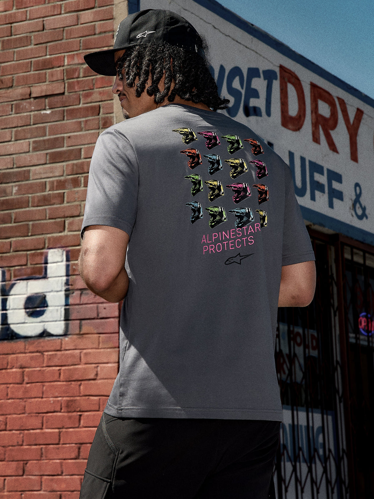 Alpinestars Pop Helmet CSF Tee, Kurzarm-T-Shirt, Anthrazitgrau, Rückengrafik mit bunten M10-Helmen und pinkfarbenem „ALPINESTAR PROTECTS“-Schriftzug, lässige Motorsport-inspirierte Streetwear