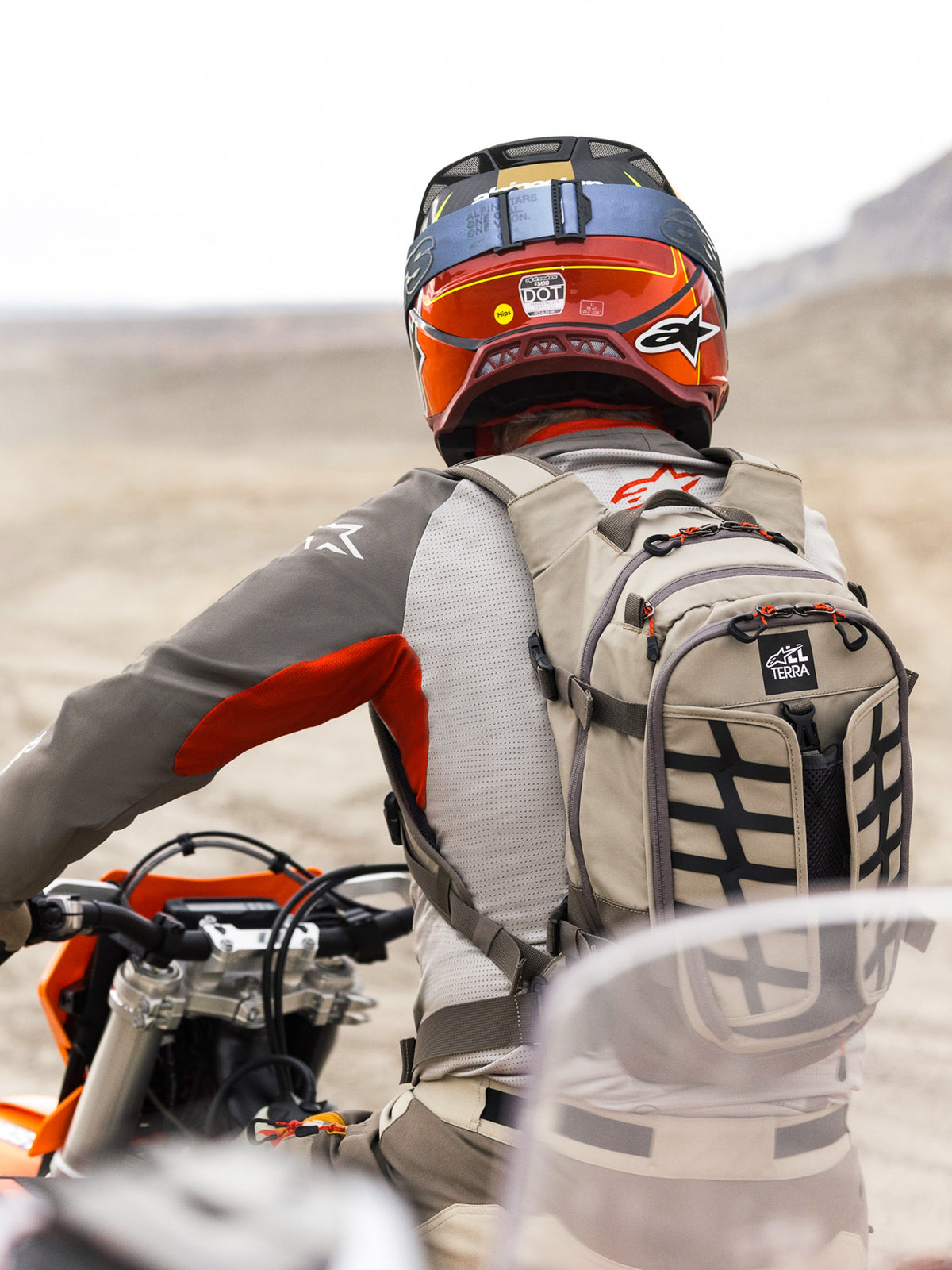 Alpinestars AT-15 Rucksack in Aluminum Walnut, getragen von einem Adventure-Motorradfahrer in einer Wüstenlandschaft. Robuste beige Textilkonstruktion mit schwarzem Gurtband, orangefarbenen Reißverschluss-Akzenten und 'All Terra'-Branding. Kombiniert mit einem rot-orangefarbenen Helm und grauer Fahrbekleidung für Offroad-Touren.