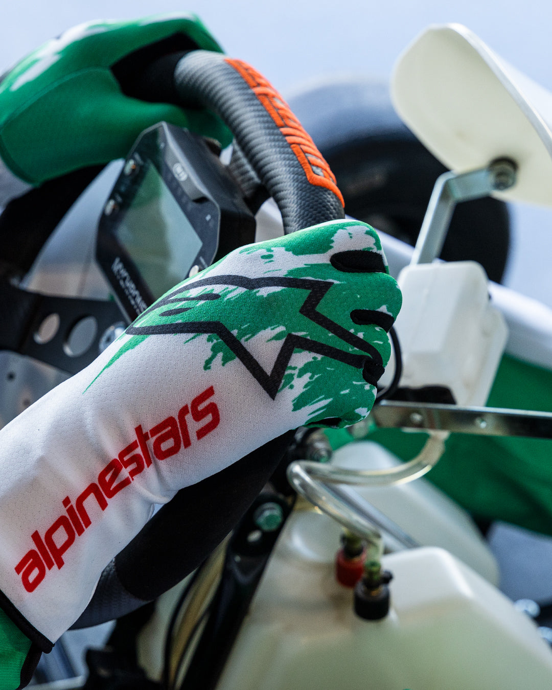 Alpinestars Tech-1 K V3 Handschuhe in Weiß, Grün und Rot, Hände eines Fahrers am Kart-Lenkrad, mit grünem sublimiertem Splash-Design, schwarzem Stern-Logo und rotem Alpinestars-Schriftzug an der Manschette, professionelle Kart-Ausrüstung.