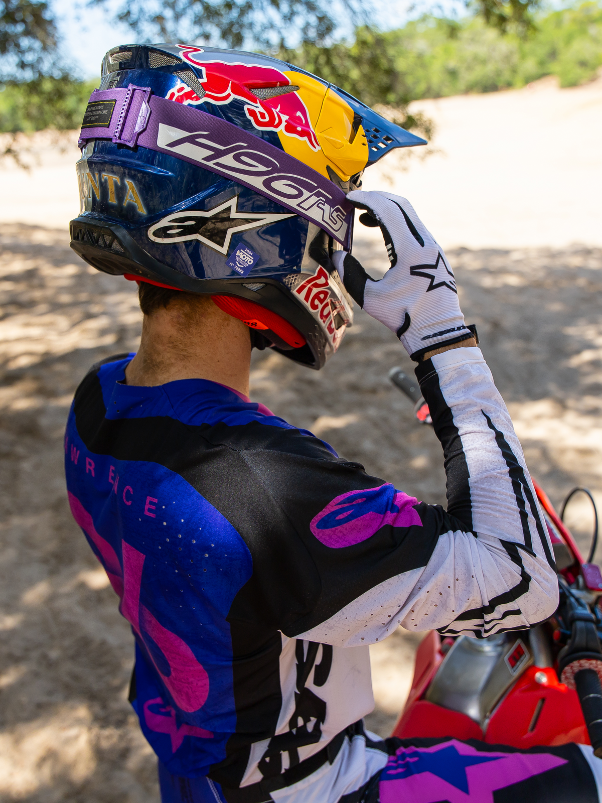 Motocross rider Hunter Lawrence wearing Limited Edition HUNTA96 XXV Supertech Vision Goggles, purple strap with 'H96' and yellow stitching, blue Red Bull helmet, purple and white jersey with black accents, white gloves, adjusting goggles in a sandy off-road setting