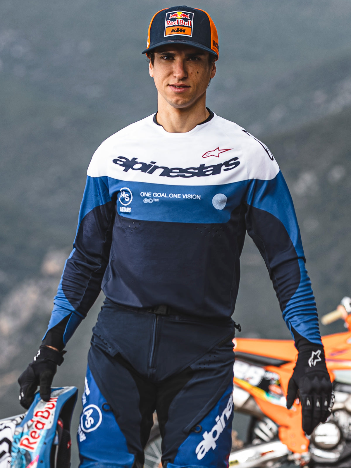 Motocross rider wearing Alpinestars Pro-Dura Jersey and Pants in Navy Bright Blue and White, featuring 'One Goal One Vision' branding, black gloves, and holding a racing helmet, professional off-road racing gear.