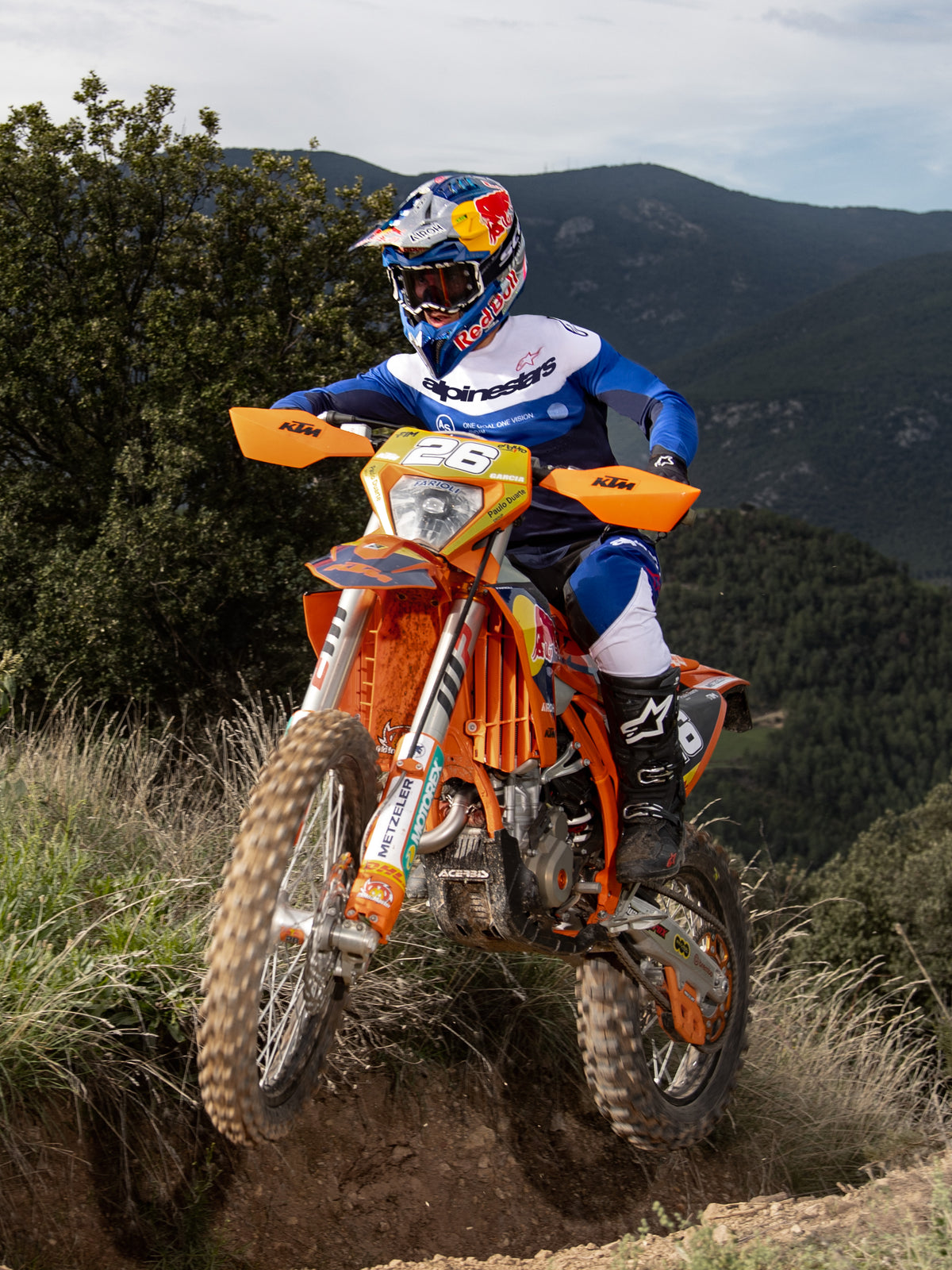Enduro rider on an orange dirt bike, Alpinestars Pro-Dura Jersey in Navy Bright Blue White, black and white riding pants, and black Alpinestars boots, navigating a rugged trail with mountains and trees in the background.
