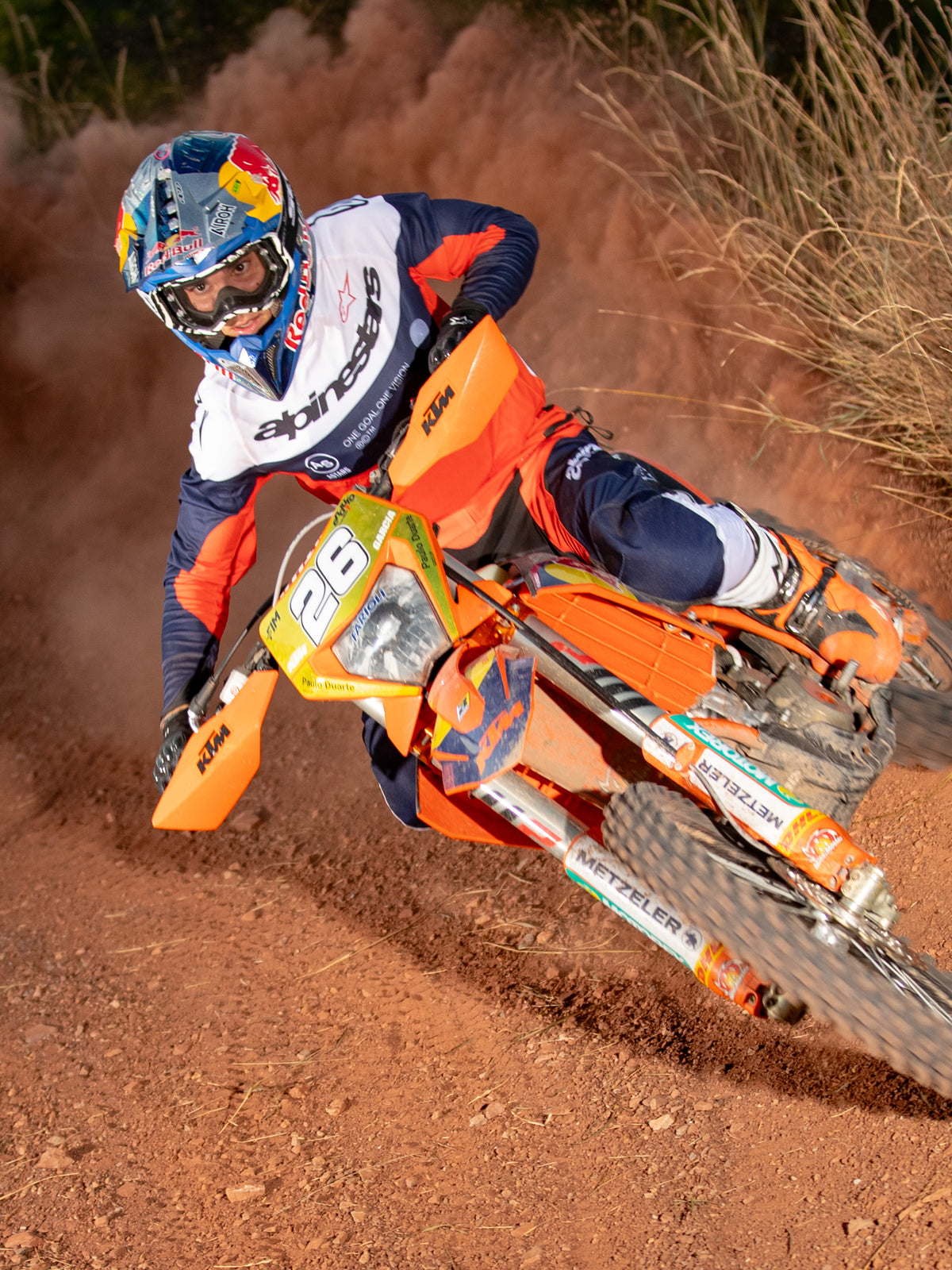 Motocross rider leaning into a turn on an orange motorcycle, wearing Alpinestars Pro-Dura Jersey and Pants in Navy, Hot Orange, and White, coordinated off-road racing gear, Red Bull branded helmet, white boots, action shot on a dusty dirt track.