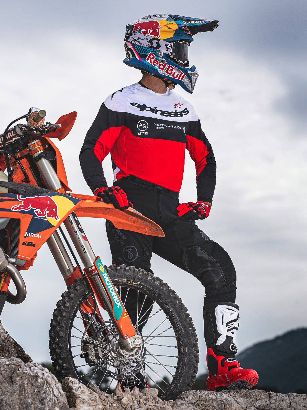 Motocross-Fahrer im Alpinestars Pro-Dura Trikot in Schwarz, leuchtendem Rot und Weiß, steht neben einem KTM Dirtbike auf felsigem Gelände, kombiniert mit einer schwarzen Hose und roten Motocross-Stiefeln, Outdoor-Enduro-Setting.