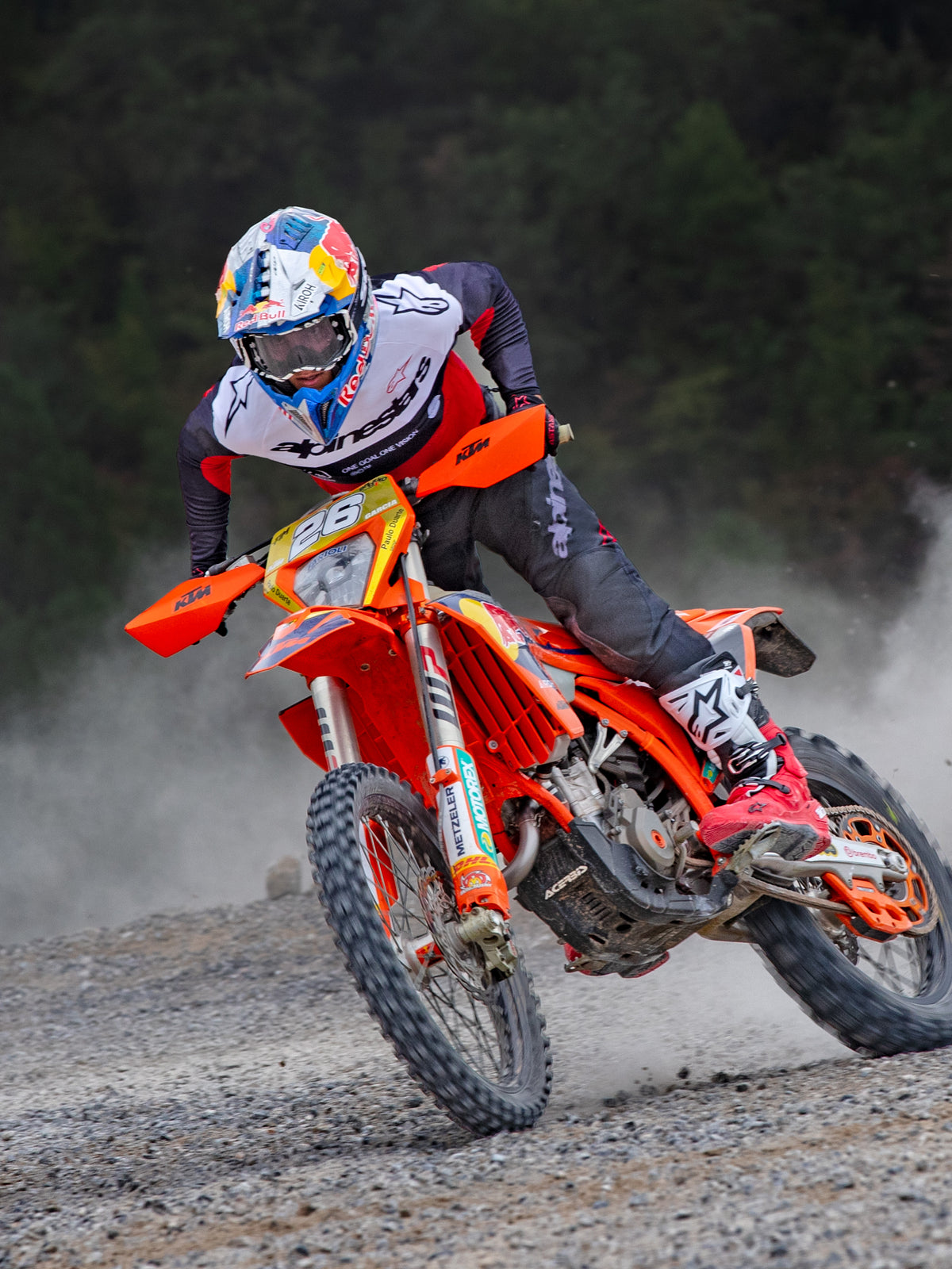 Motocross-Fahrer in Aktion im Alpinestars Pro-Dura Jersey in Schwarz, leuchtendem Rot und Weiß, fährt ein orangefarbenes Dirtbike auf einem staubigen Offroad-Trail, kombiniert mit einer schwarzen Hose und rot-weißen Stiefeln