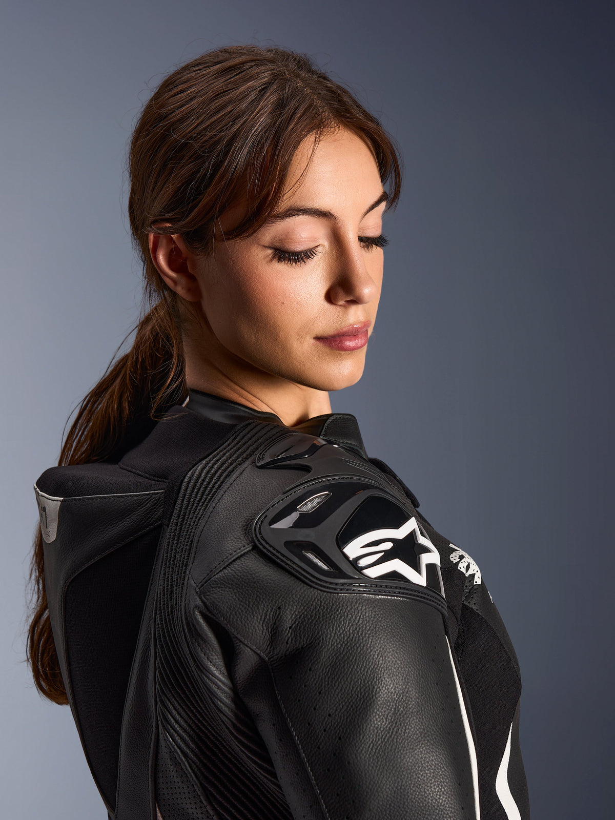 Frau im Alpinestars Stella GP-R7 2-Teiler-Lederanzug, Nahaufnahme der schwarzen Lederschulter mit DFS RR Panzerung und weißem Stern-Logo, mit integriertem Aero-Höcker und perforierten Details.